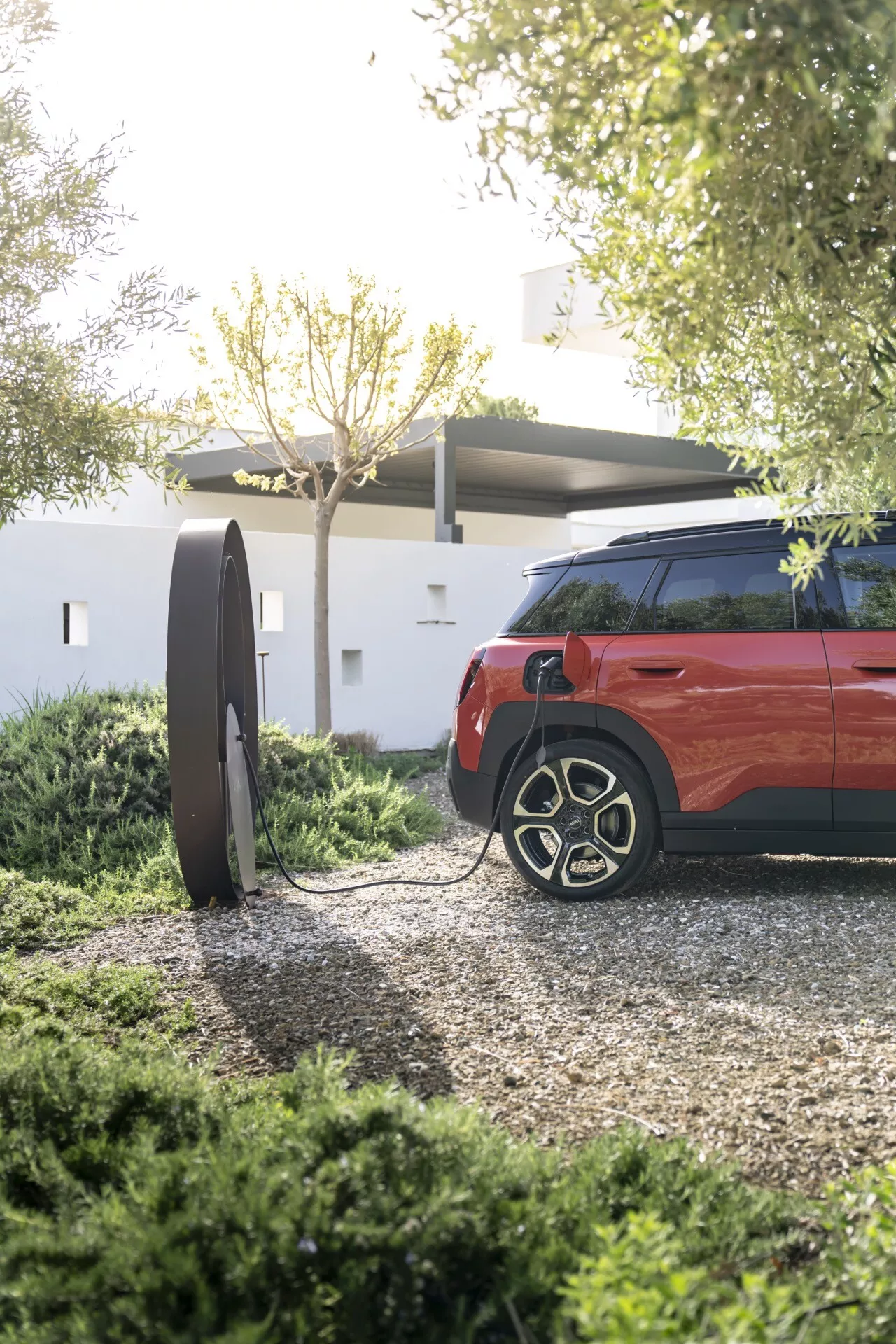 2024 Mini Aceman (J05) SUV in vivid red, highlighting the car's rear quarter and wheel design with an electric vehicle charging station in view, suggesting sustainable transportation technology.