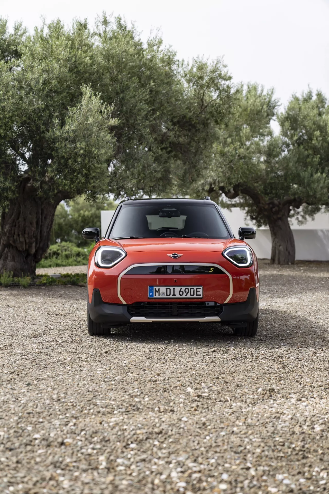 2024 Mini Aceman (J05) in red, showcasing distinctive Union Jack LED tail lights, rear spoiler, parking sensors, and electric model indication from a direct rear view on gravel with olive trees and a building in the background