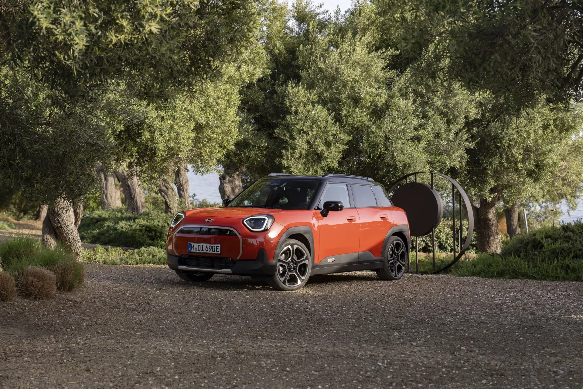 2024 Mini Aceman (J05) in a three-quarter front view on a gravel path with greenery, showcasing its LED headlights, multi-spoke alloy wheels, two-tone body color with black roof, sporty hood design, and compact SUV silhouette