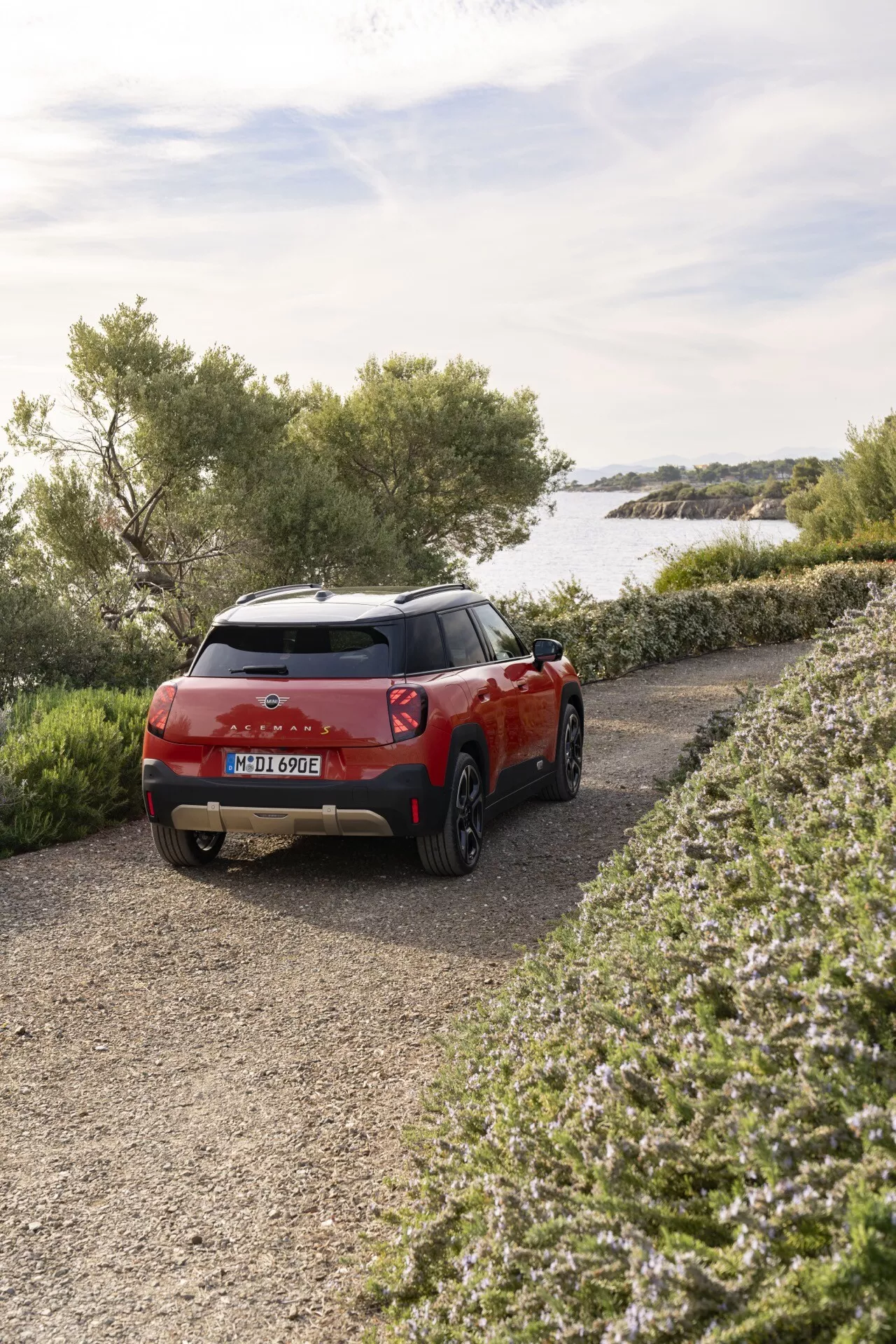 2024 Mini Aceman compact SUV in red, showcasing rear three-quarter angle view near a lake or calm sea, highlighting rear lights and contour, surrounded by natural scenery on a gravel path, indicating off-road capabilities and exterior design features