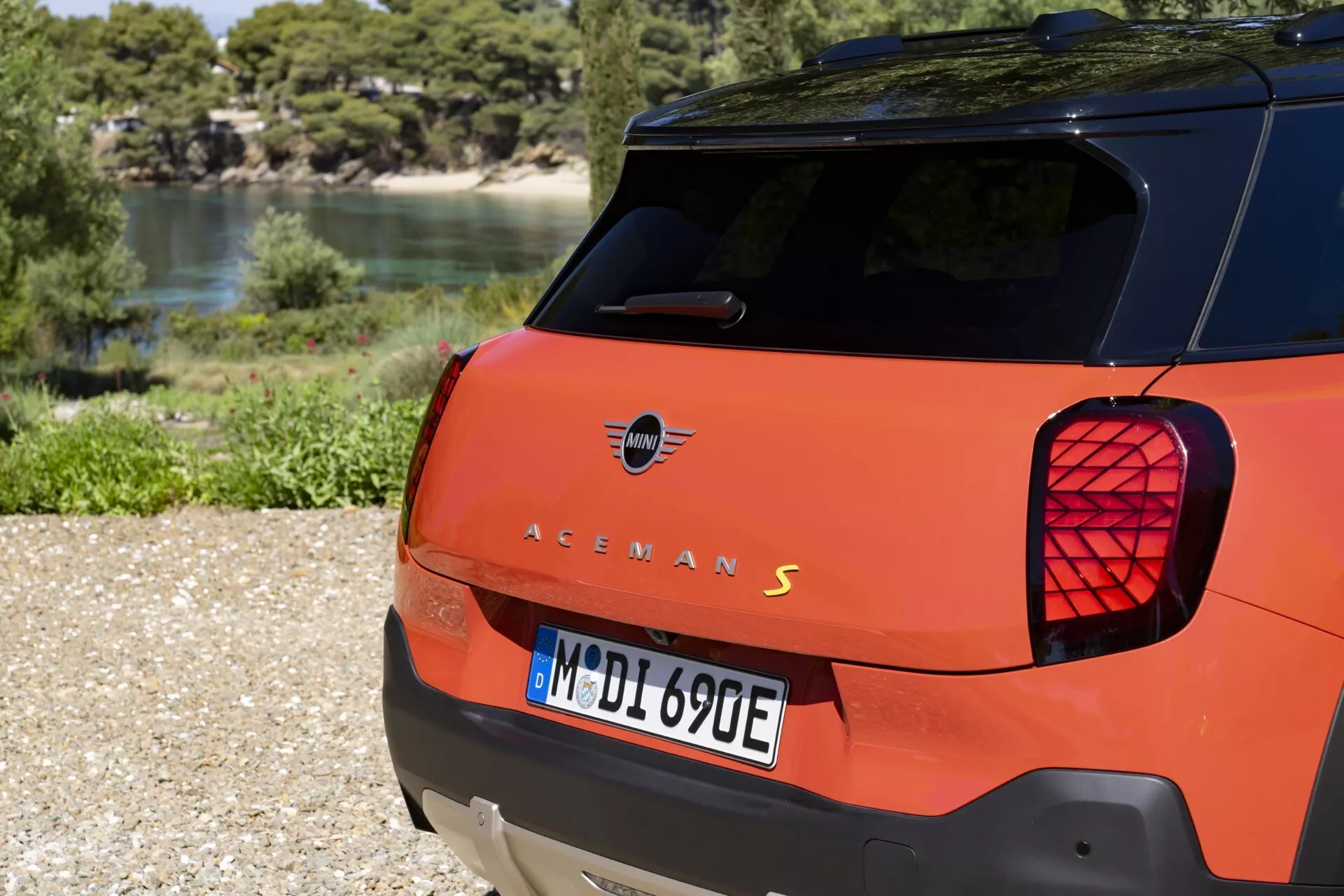 Rear view of an orange hatchback showing distinctive Union Jack motif tail lights, possibly electric 'S' model, parked outdoors on gravel with natural background