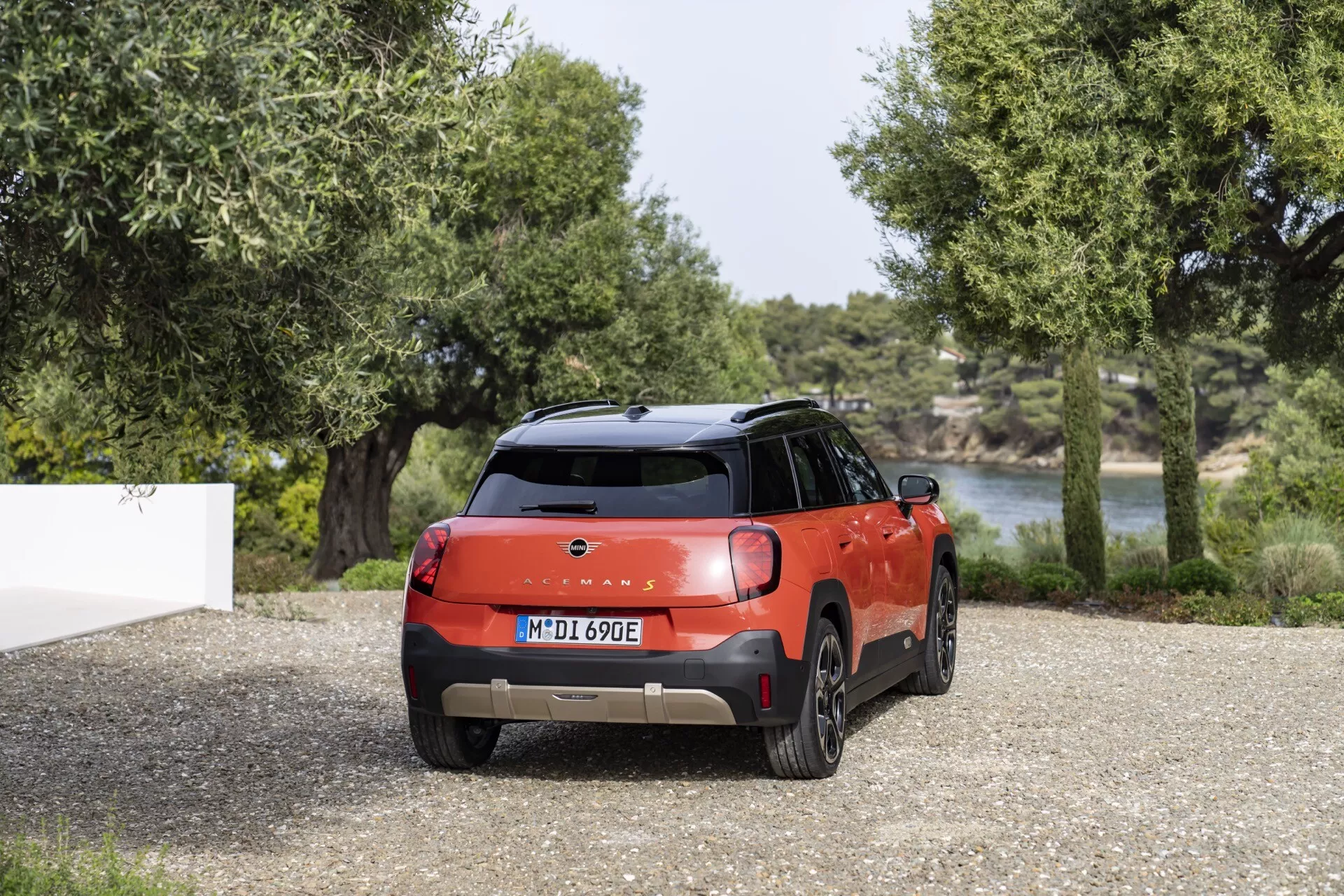 2024 Mini Aceman (J05) rear view highlighting its two-toned color scheme, sleek LED taillights, and sporty rear spoiler against a natural backdrop with gravel, trees, and water hinting at electric or hybrid capability