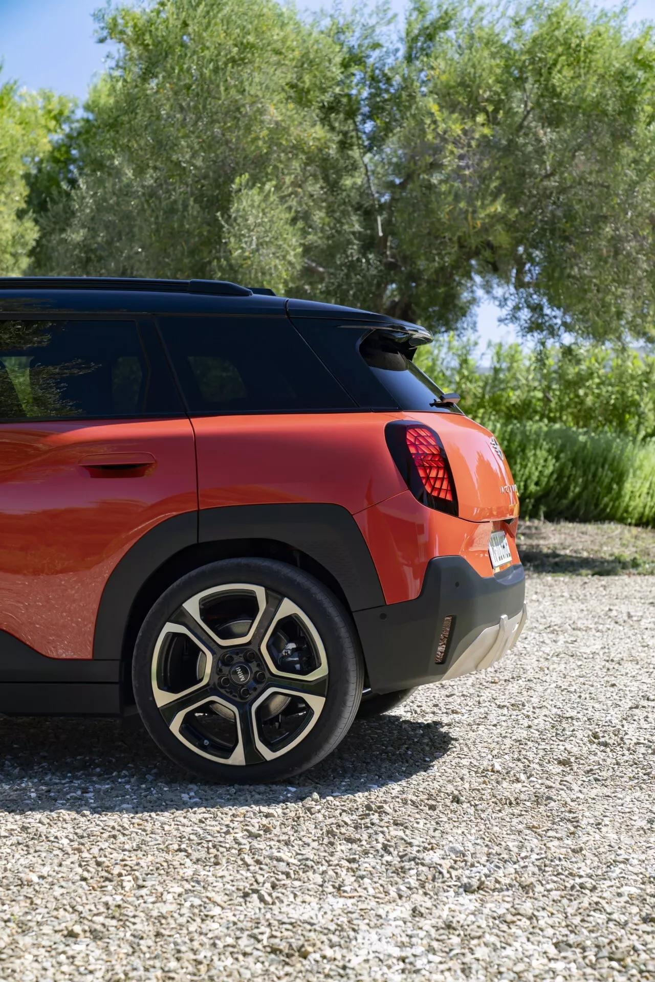 Three-quarter rear view of the 2024 Mini Aceman (J05) showcasing its right side from an elevated angle, featuring a vibrant red-orange matte finish, five-double-spoke alloy wheels, LED taillight configuration, rear bumper with parking sensors, and a rear windshield wiper, set against an outdoor backdrop with greenery.