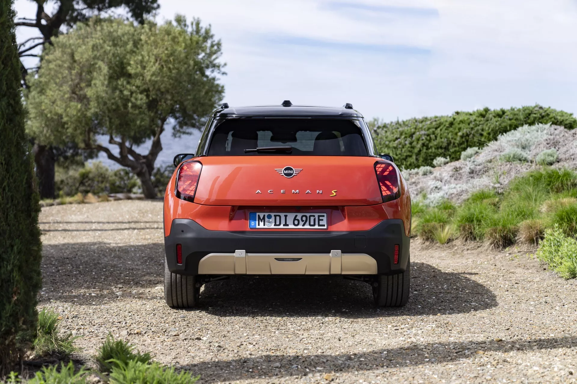 Rear view of the 2024 Mini Aceman (JO5) parked on a gravel path featuring emblem, model name with 'E' designation, contrasting rear bumper, LED tail lights, and roof rails surrounded by natural scenery