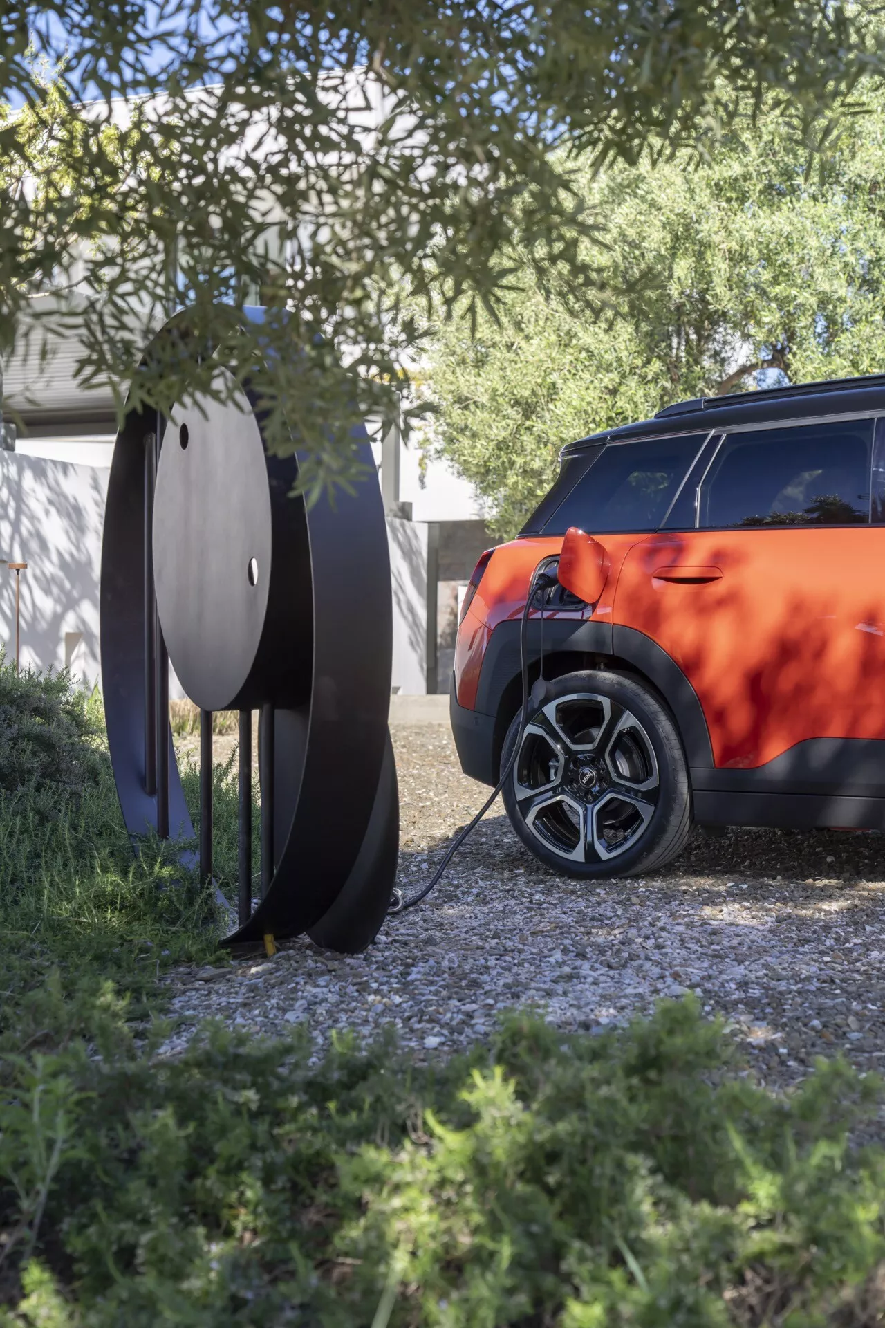 Partial view of a 2024 Mini Aceman (JO5) featuring the rear-right side with a multi-spoke alloy wheel, vibrant bodywork, and modern tail light design, framed by natural foliage and a circular outdoor sculpture, under natural daylight