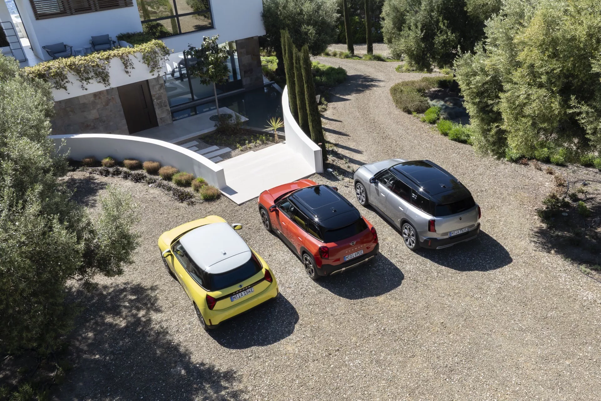 Aerial view of three 2024 Mini Aceman (J05) models in yellow, red, and silver parked on a gravel driveway next to a modern residence, highlighting their compact hatchback, sedan, and SUV body styles