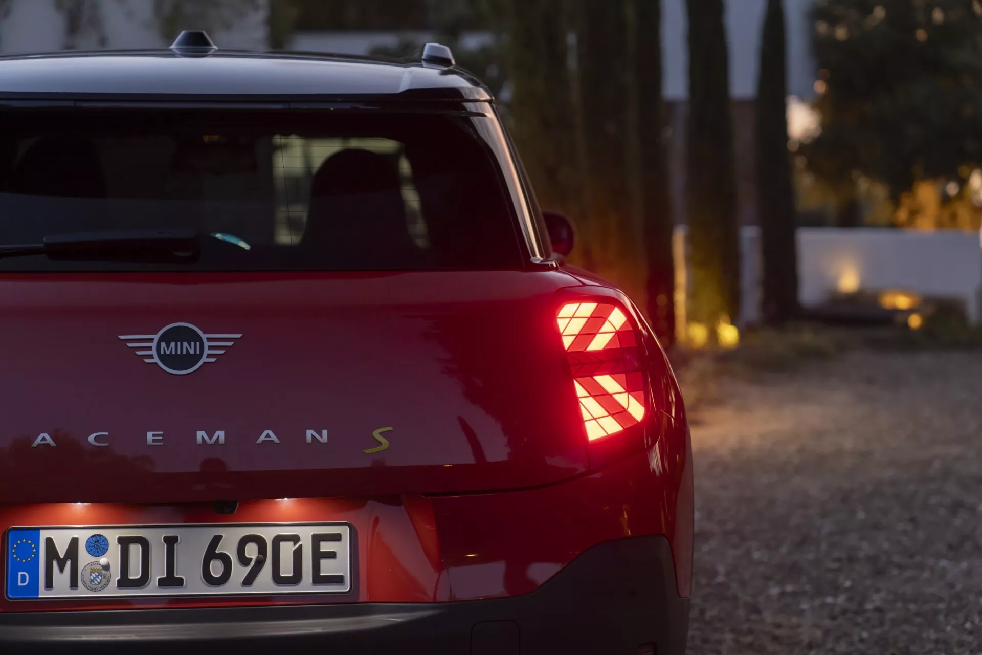 2024 MINI Aceman S viewed from a low rear three-quarter perspective highlighting its unique LED taillights with a Union Jack pattern, dark red body with contrasting black roof, and eco-friendly European license plate, in a serene twilight setting