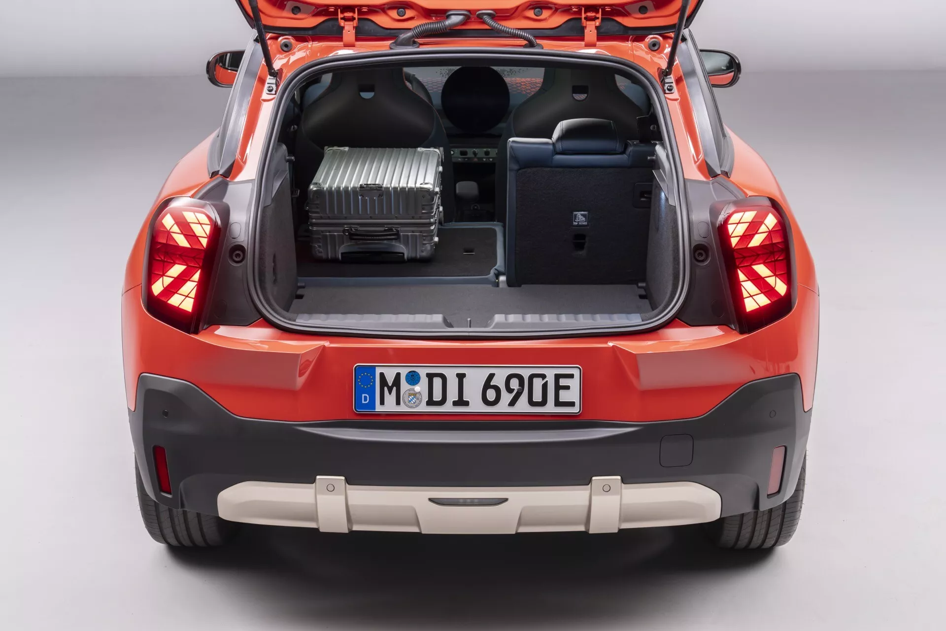 Rear-view image of 2024 Mini Aceman (J05) with hatchback open showing trunk space and interior glimpse, featuring LED tail lights, license plate, rear bumper with parking sensors, dual-tone bumper design, visible electric powertrain component, and hatchback interior panel with grab handle
