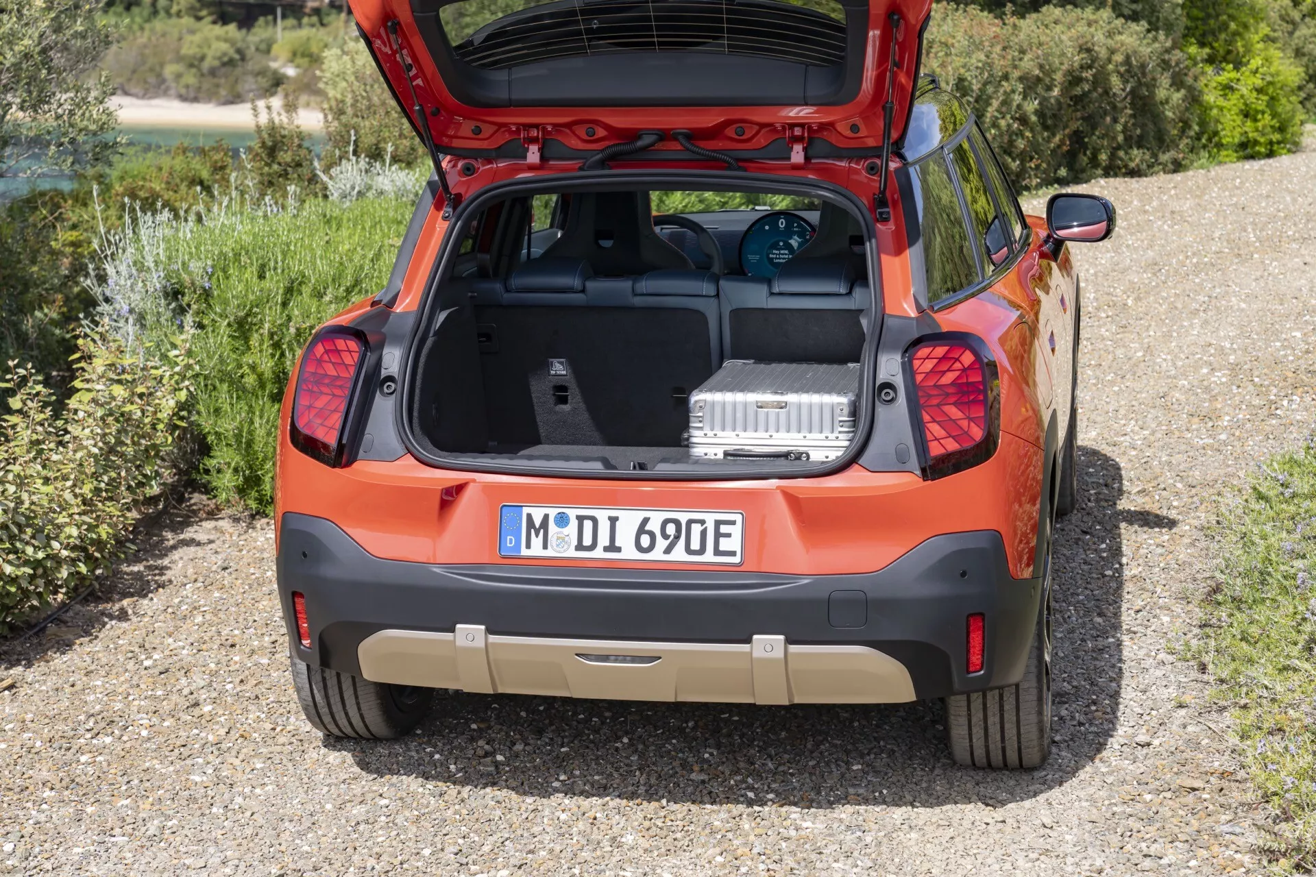 Rear view of a 2024 Mini Aceman (J05) orange compact SUV parked on gravel with the hatchback open, displaying the cargo area and featuring parking sensors, unique taillights, aftermarket trunk mat, '690E' model badging, and rear windshield wiper, against a natural landscape