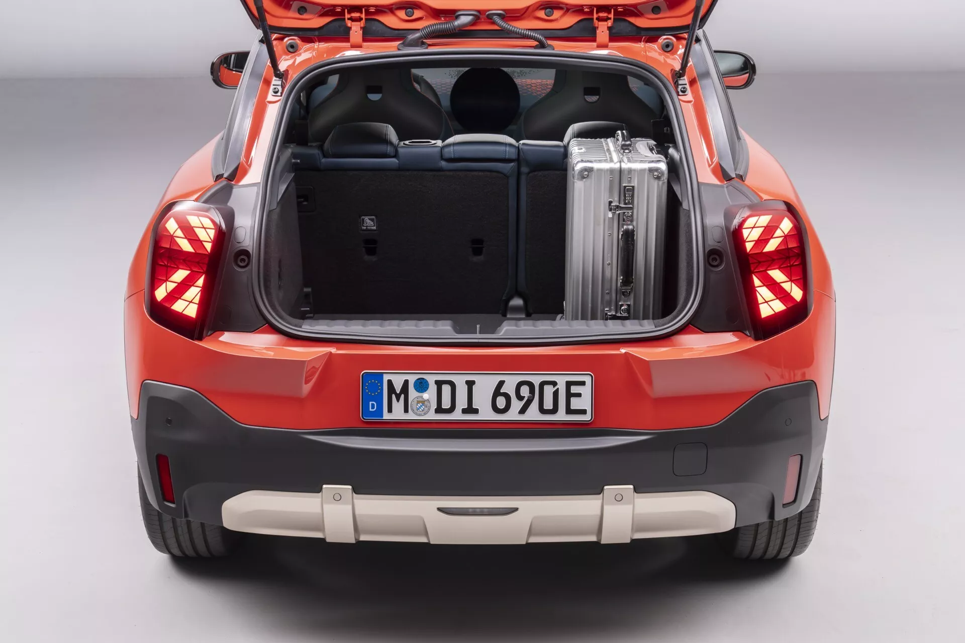 2024 MINI Aceman (JO5) rear view showcasing the spacious trunk with metallic luggage, intricate LED tail lights, parking sensors on the bumper, and vibrant orange-red color finish