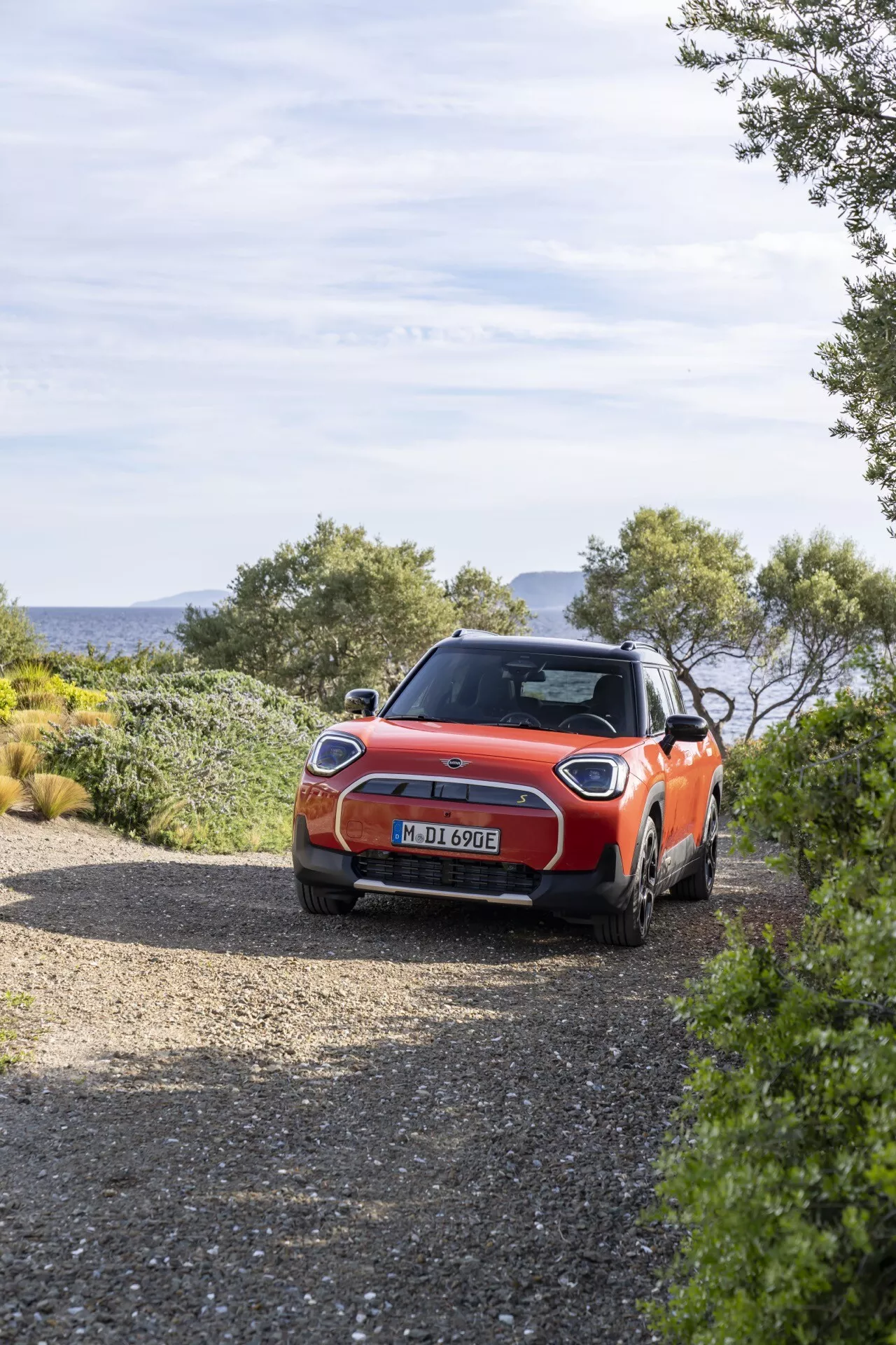 2024 Mini Aceman (J05) parked on a gravel road, featuring LED tail lights, vibrant orange glossy finish, alloy wheels, tinted rear window, roof rails, shark-fin antenna, hatchback body style, surrounded by natural scenery with potential off-road capabilities, in natural daylight