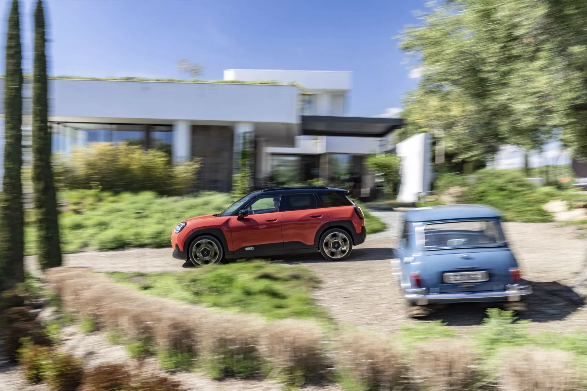 Red 2024 Mini Aceman (J05) in motion captured from a side profile, highlighting its sleek aerodynamic design and large alloy wheels, suggesting off-road capability, against a blurred residential and green landscape background.