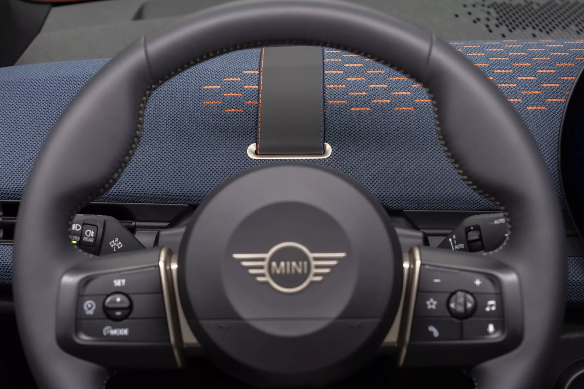 Close-up view of the lower three-quarters of a MINI Aceman 2024 steering wheel featuring logo, control buttons for media and cruise control, sporty design with thumb rests, and a partial view of a digital dashboard.