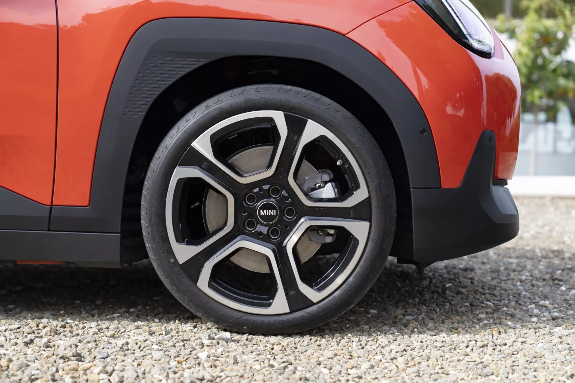 Close-up view of MINI Aceman 2024 front right wheel and lower fender in vibrant orange with five-spoke dual-tone alloy and MINI logo, showcasing tire tread and black wheel arch trim on a gravel surface.