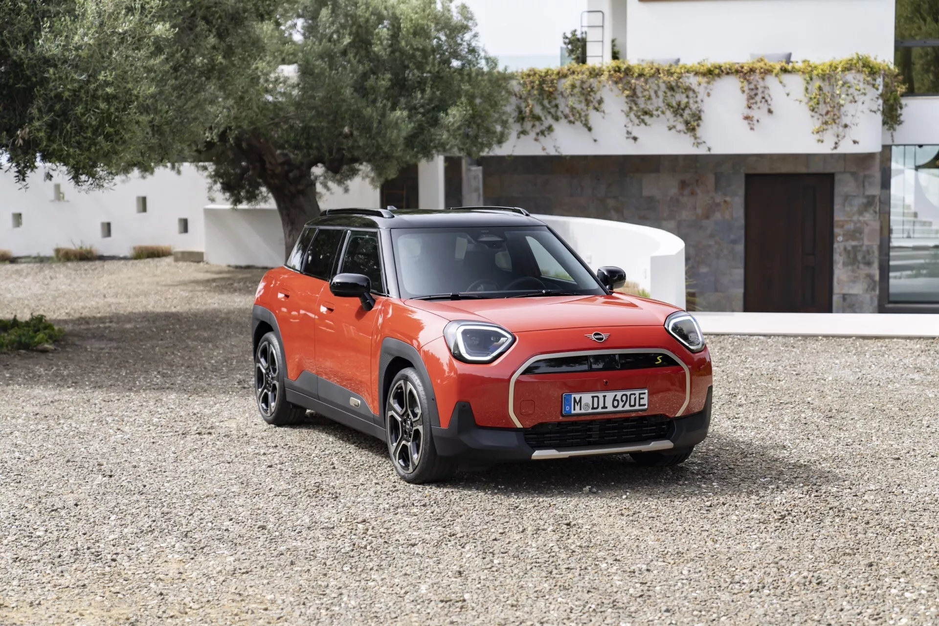 2024 Mini Aceman (JO5) in bright orange and black parked outdoors on gravel, showcasing its electric compact hatchback design with LED headlights, closed grille, multi-spoke alloy wheels, and charging port