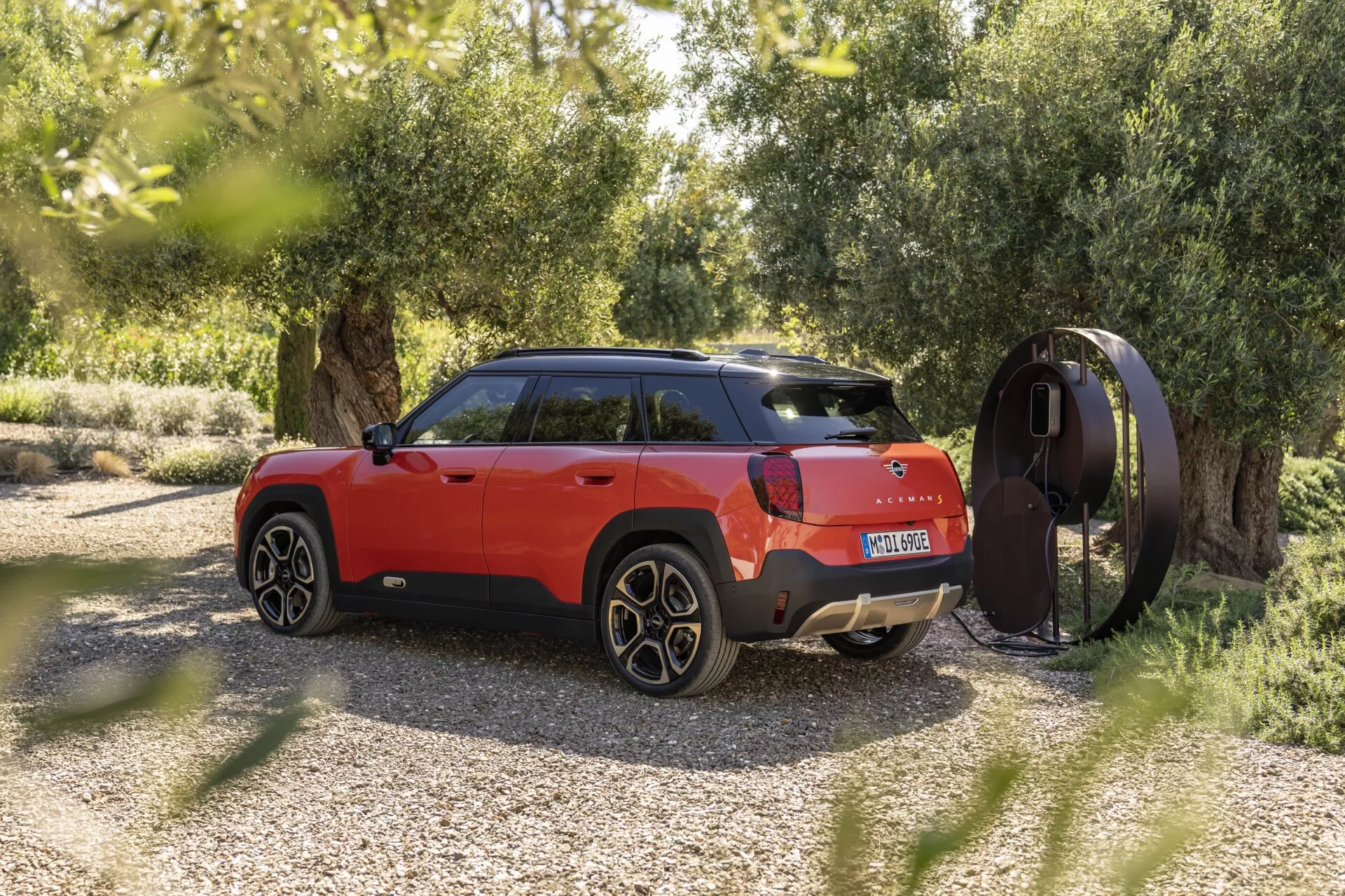 2024 Mini Aceman (J05) in vibrant red showcasing its sleek side profile and partial rear view with distinctive design elements, parked outdoors amidst greenery with a nearby charging station, highlighting its potential as an electric or plug-in hybrid SUV.
