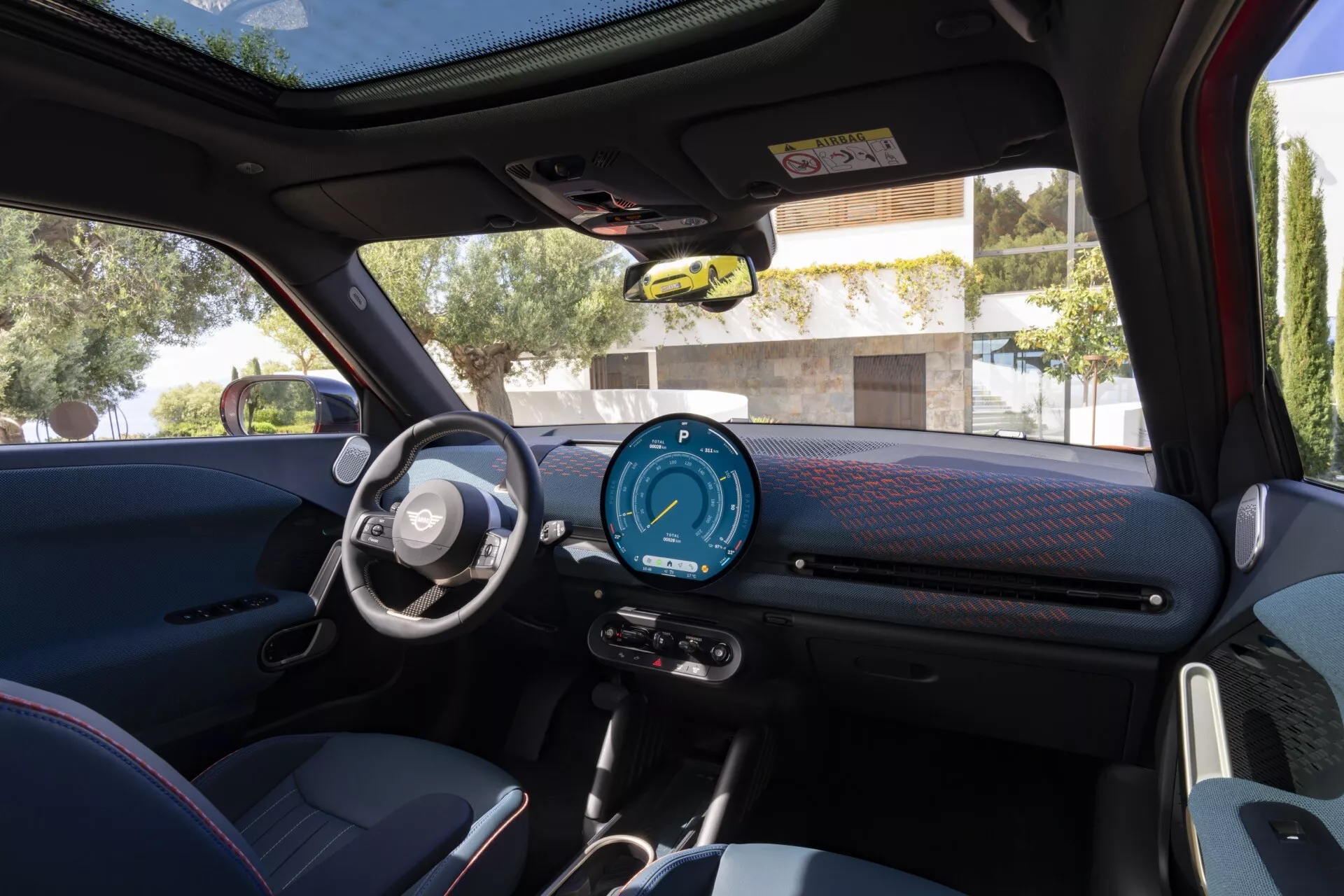 Interior view of 2024 Mini Aceman (J05) showcasing dashboard, digital instrument cluster, steering wheel with multimedia and cruise control buttons, power window and side mirror controls on driver's door, sporty textured materials, and panoramic sunroof in daylight