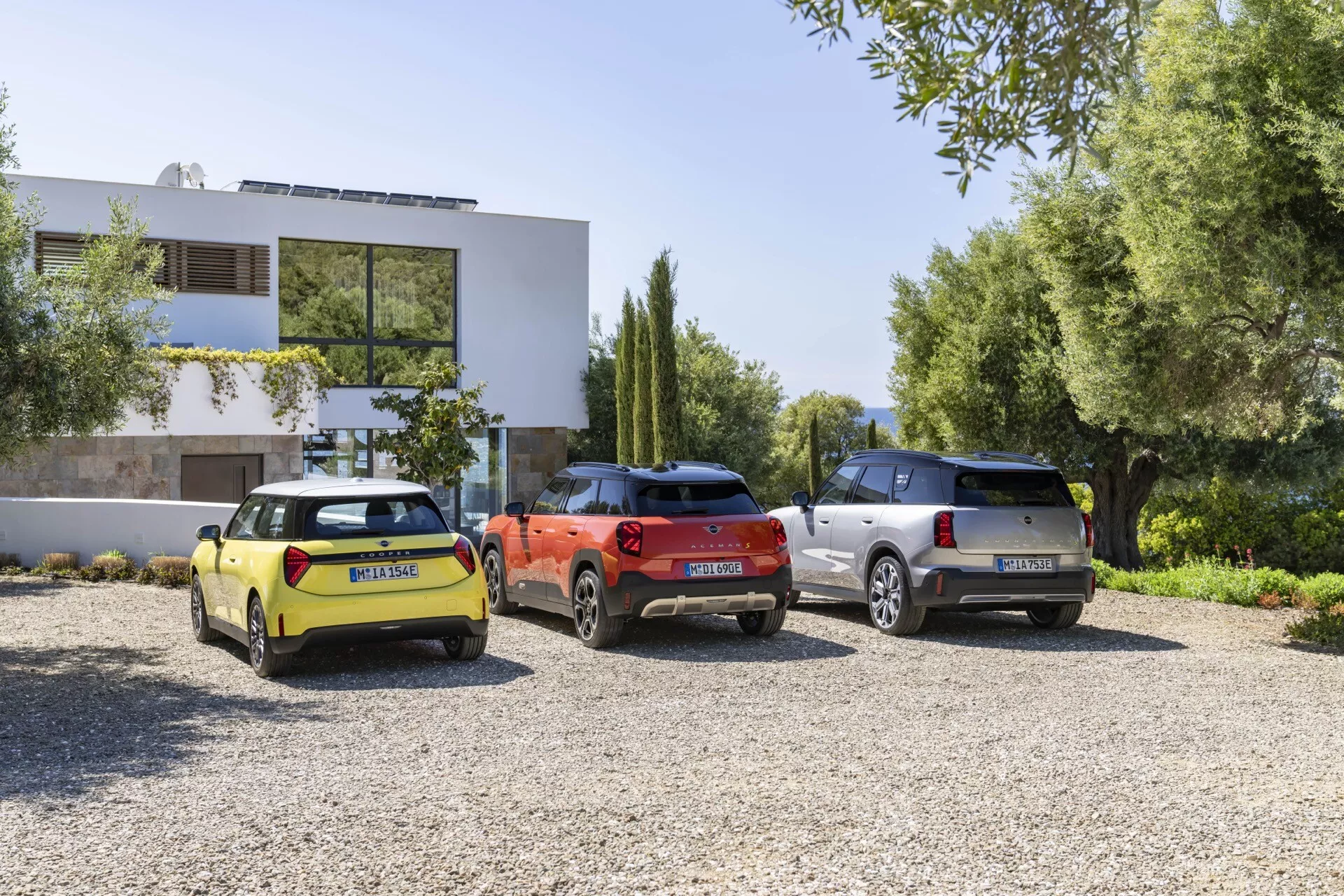 2024 Mini Aceman (J05) models in vibrant yellow, orangish-red, and silver parked on gravel in front of a modern residential building, showcasing rear three-quarter views with focus on contemporary design elements like aerodynamic lines and modern lighting
