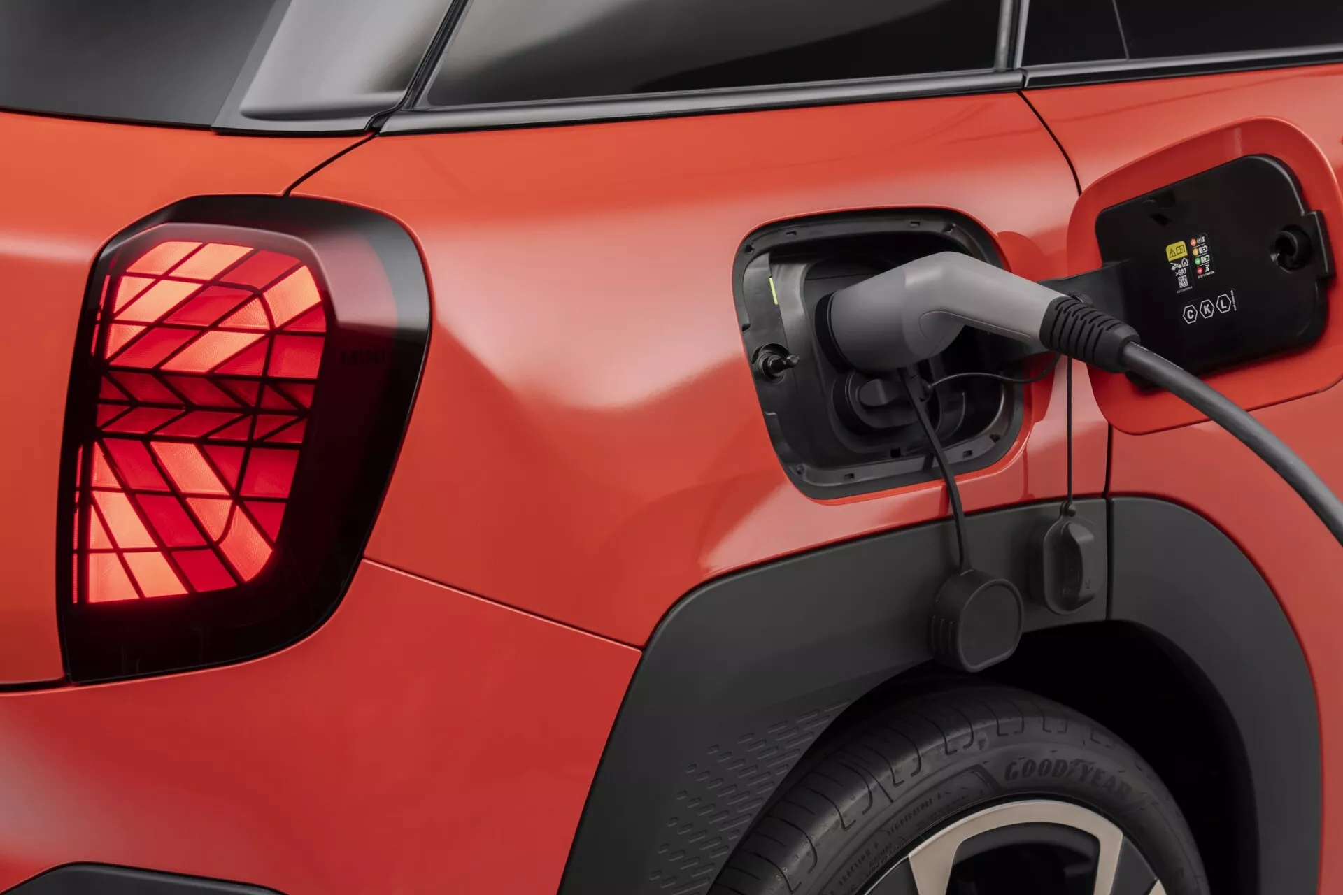 2024 Mini Aceman (J05) red electric vehicle being charged, showing close-up of open charging port with connected cable, LED tail lights, and rear wheel