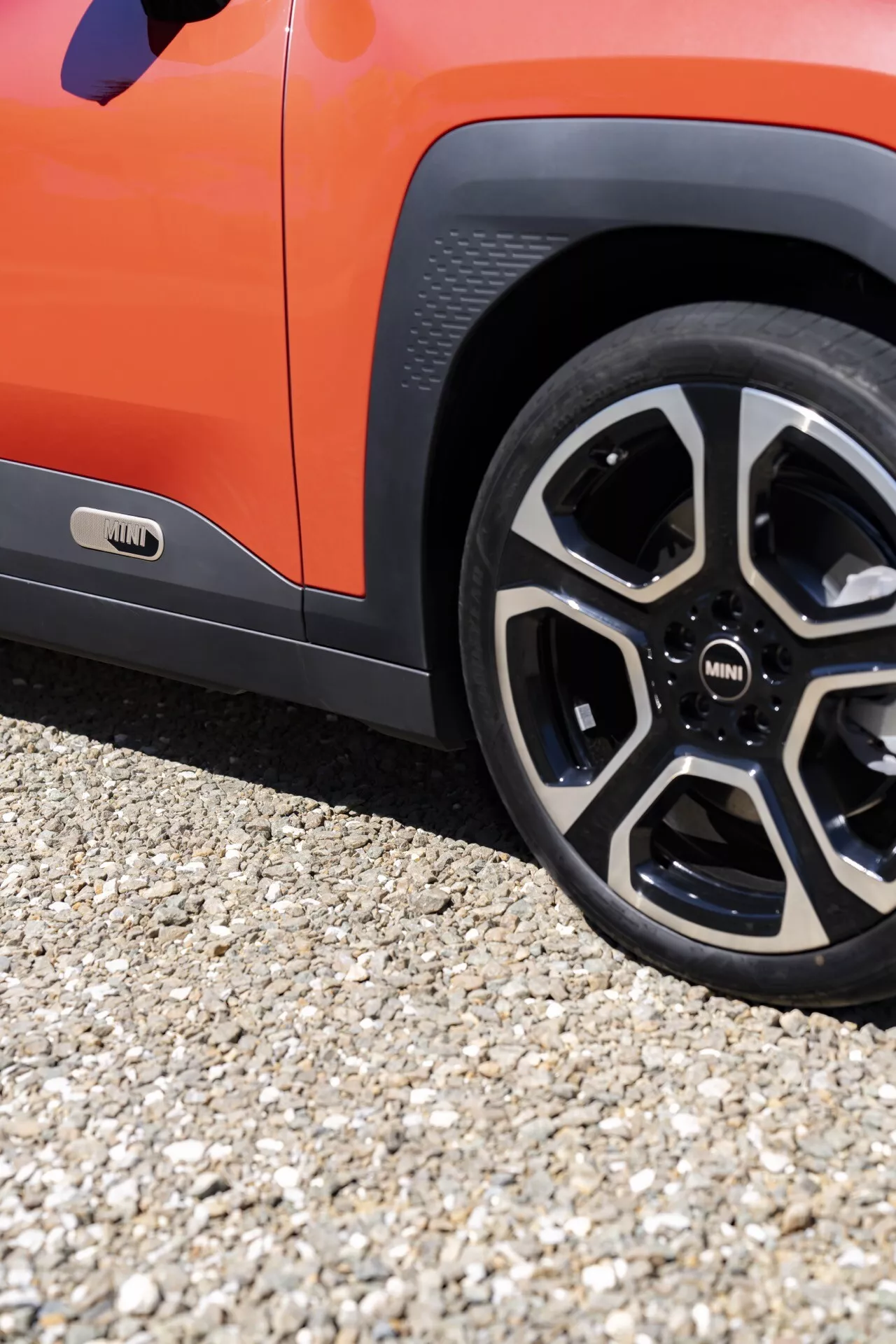 Detailed view of the Mini Aceman 2024 (JO5) focusing on the lower side profile, including a close-up of the rear wheel with a modern multi-spoke design and a portion of the orange front door, highlighting features like the side sill badge, possibly indicating an electric or hybrid model, parked on a gravel surface