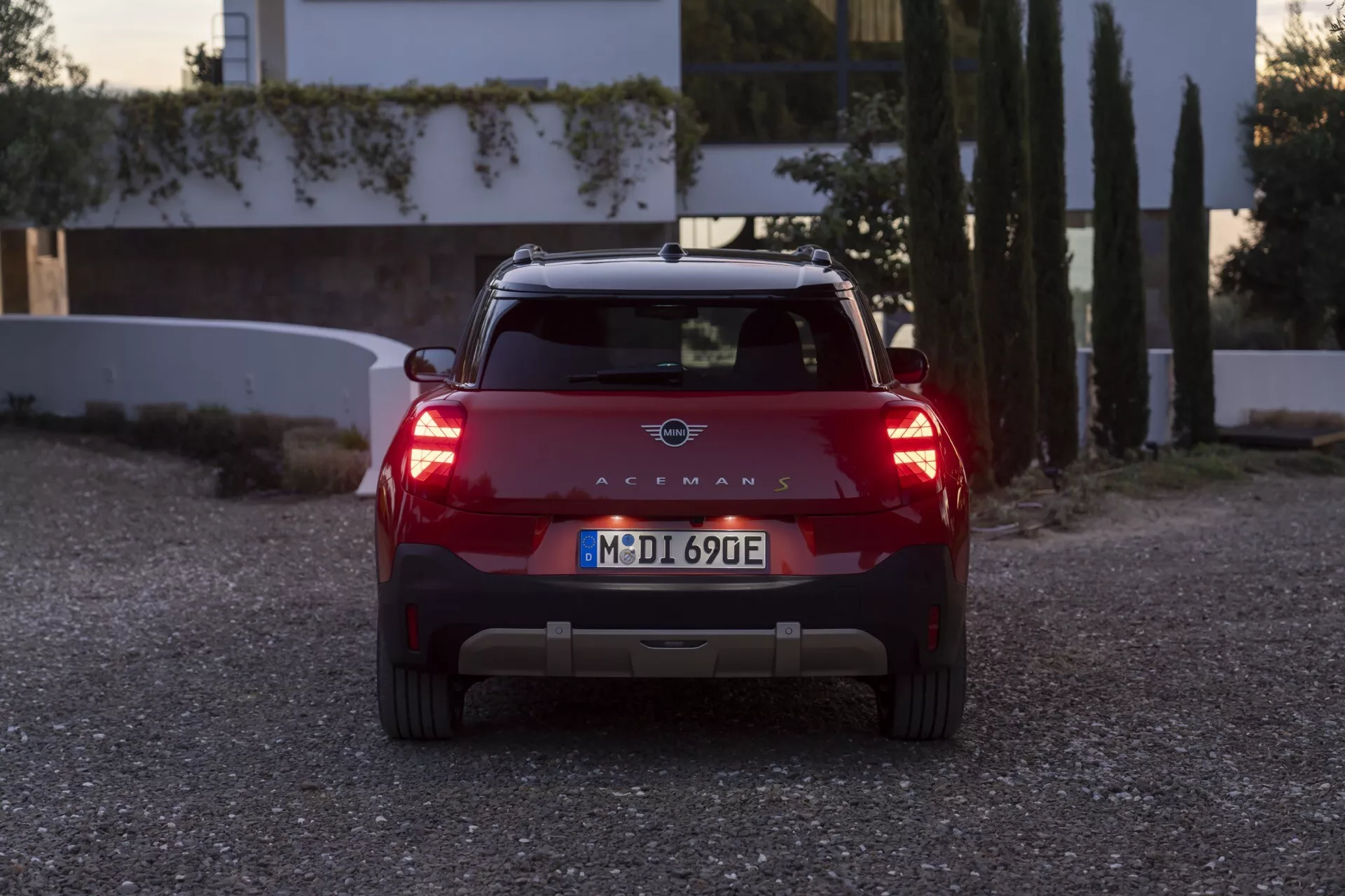 Rear view of the 2024 Mini Aceman (J05) compact SUV in red at dawn or dusk with lights on, showcasing distinct LED taillights, brand emblem, European license plate, rear spoiler with integrated brake light, parking sensors, and potentially electric vehicle indications without visible exhaust, against a residential background.