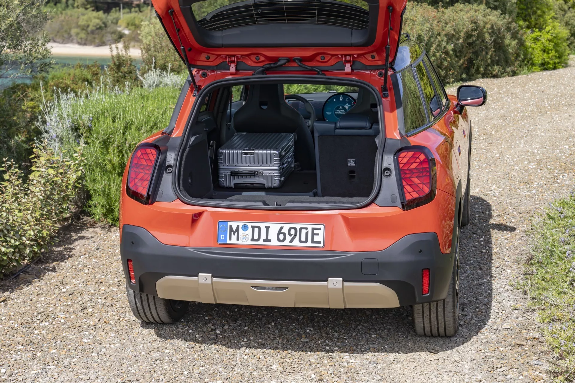 2024 Mini Aceman (J05) orange hatchback rear view with open trunk showing EV charging device, parked on gravel surrounded by greenery