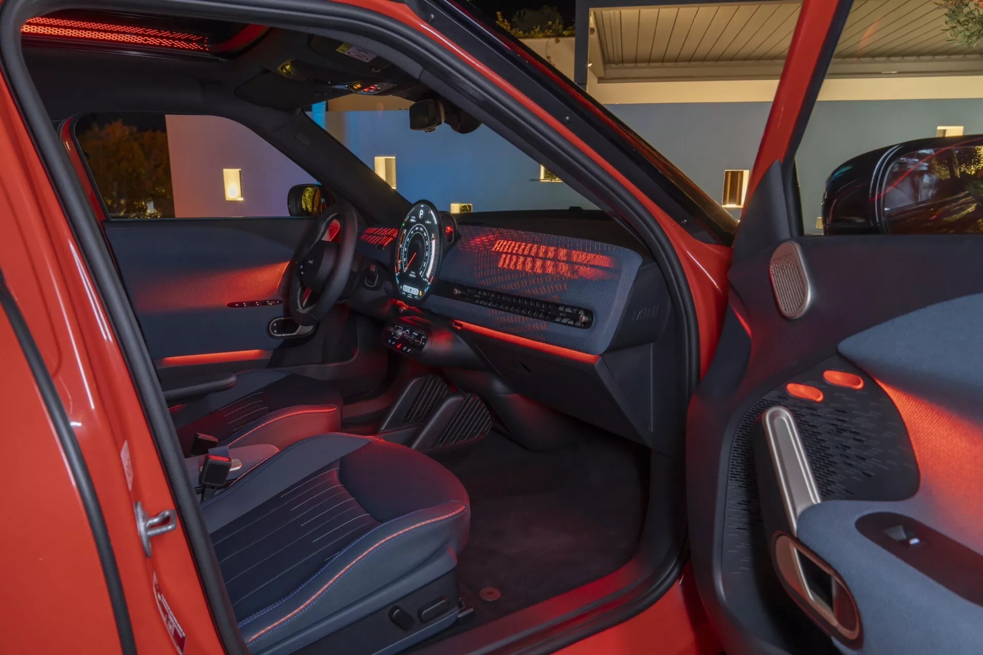 Interior view of the Mini Aceman 2024 (J05) from driver's side with open door, showing illuminated dashboard, infotainment display, multifunction steering wheel, high-quality fabric seats, and modern cabin features