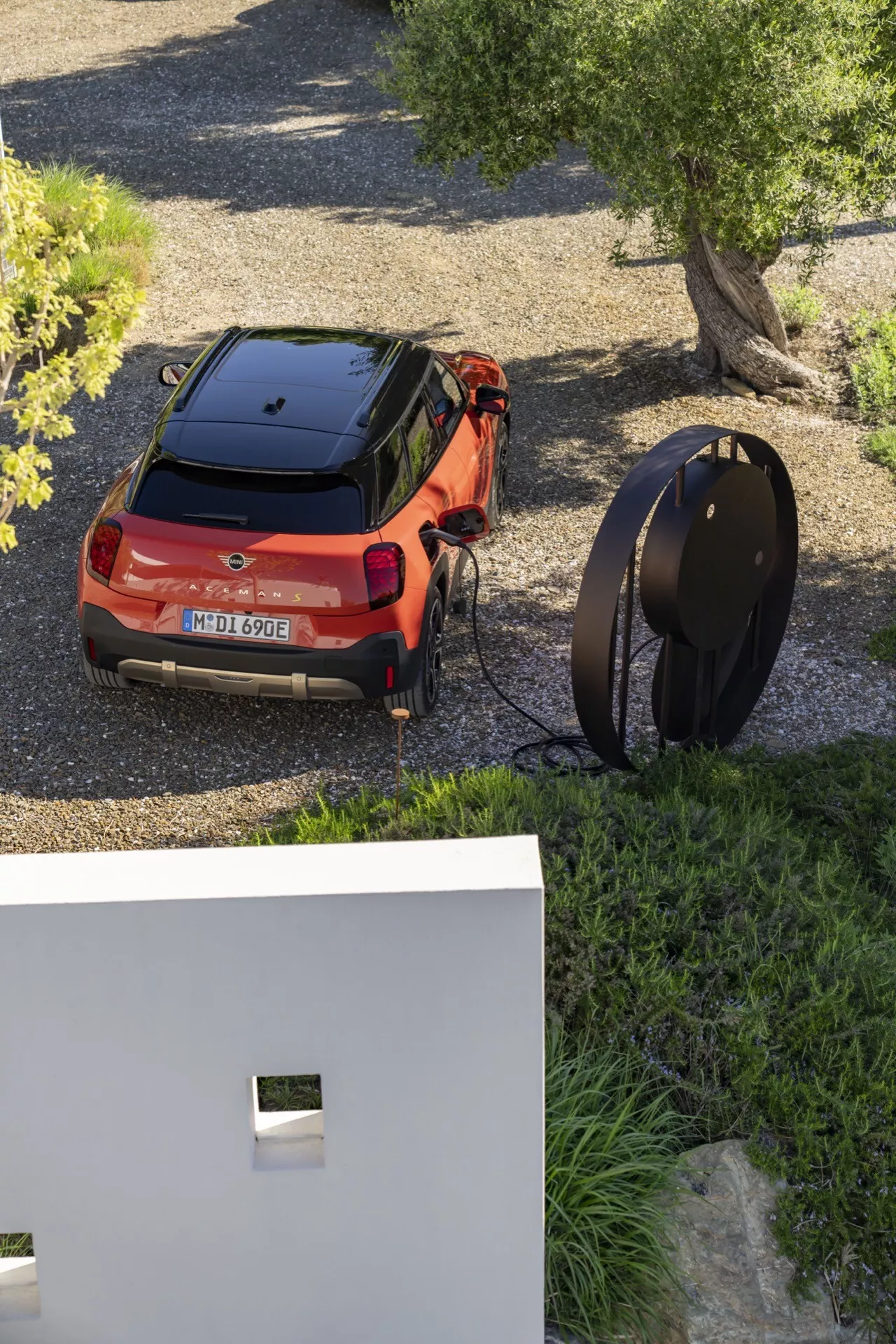 High-angle view of an orange 2024 Mini Aceman (J05) SUV parked and charging, showcasing its contemporary design with a two-tone color scheme, slim tail lights, and electric charging capability, set against a modern residential backdrop