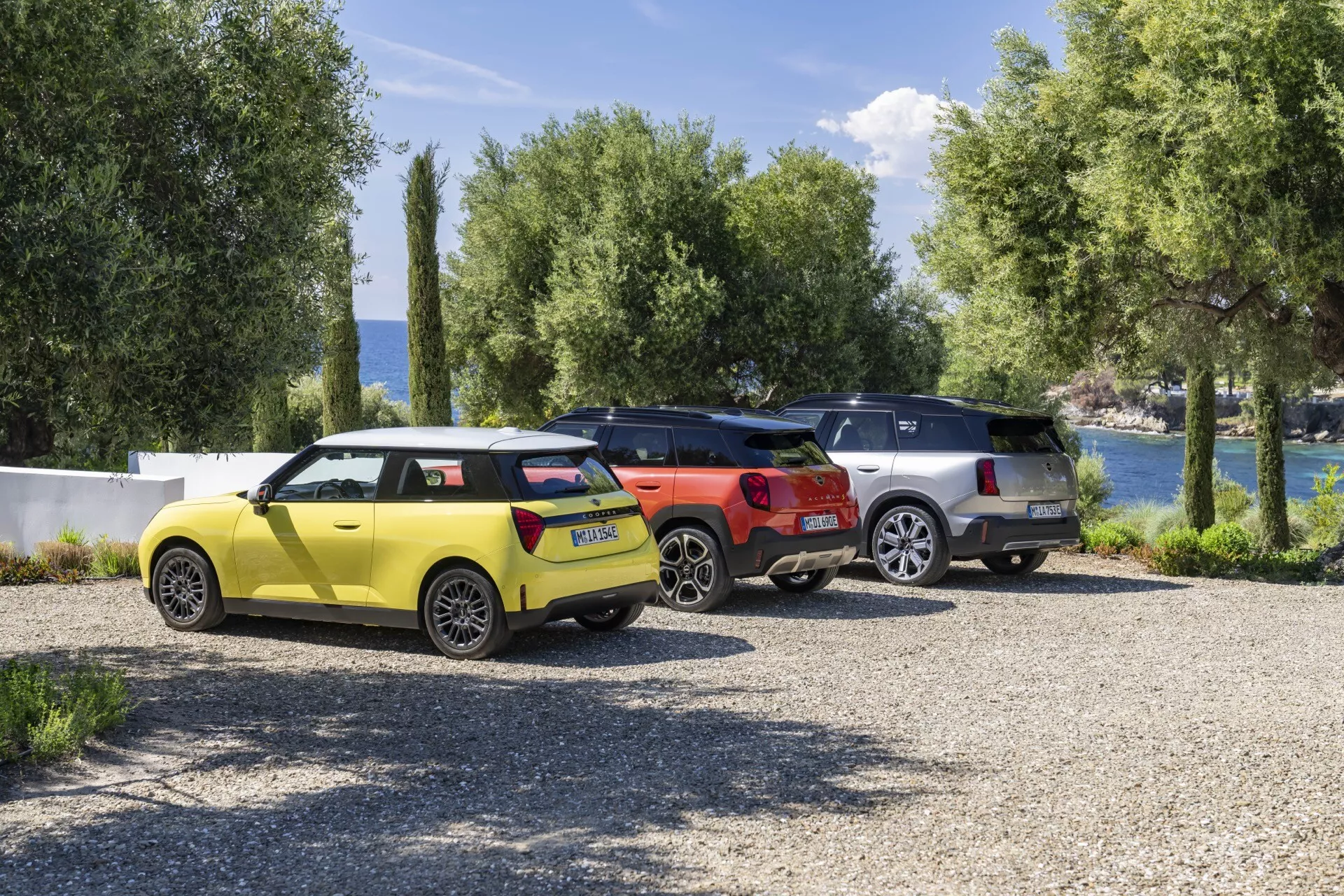 2024 Mini Aceman (J05) in vibrant yellow with modern hatchback design parked alongside a sleek red SUV and a sophisticated silver crossover against a picturesque lakeside scene with lush foliage, encapsulating a leisurely vacation atmosphere