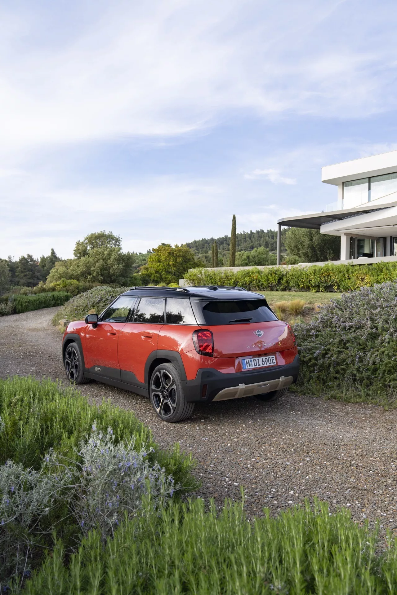 2024 Mini Aceman (J05) in red with black details, modern SUV on gravel with greenery, showcasing rear and side design highlights under clear daylight