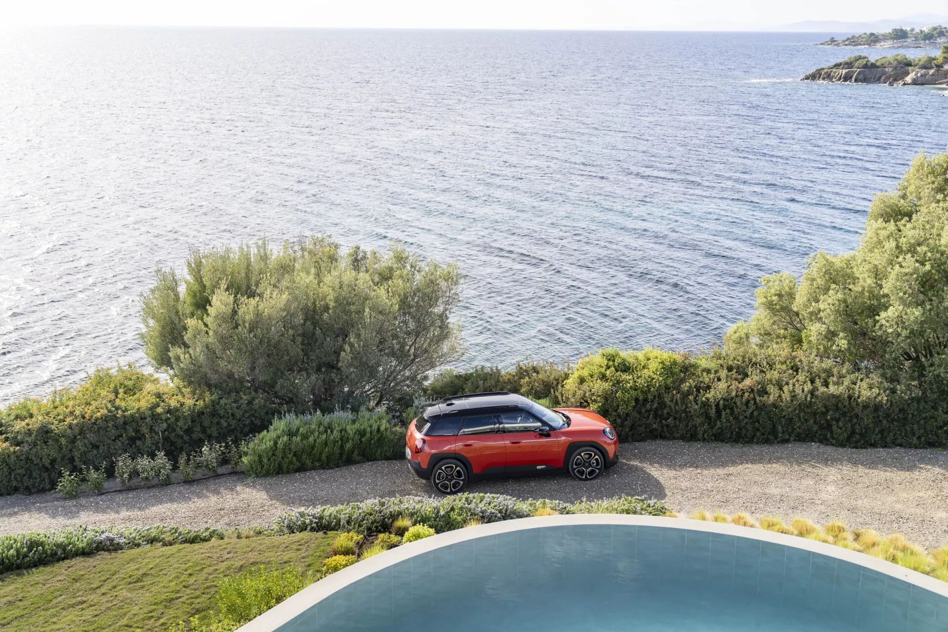 2024 Mini Aceman (J05) in vivid red parked by the sea near a manicured garden and swimming pool, showcasing its compact SUV design and modern aesthetics from a high-angle view