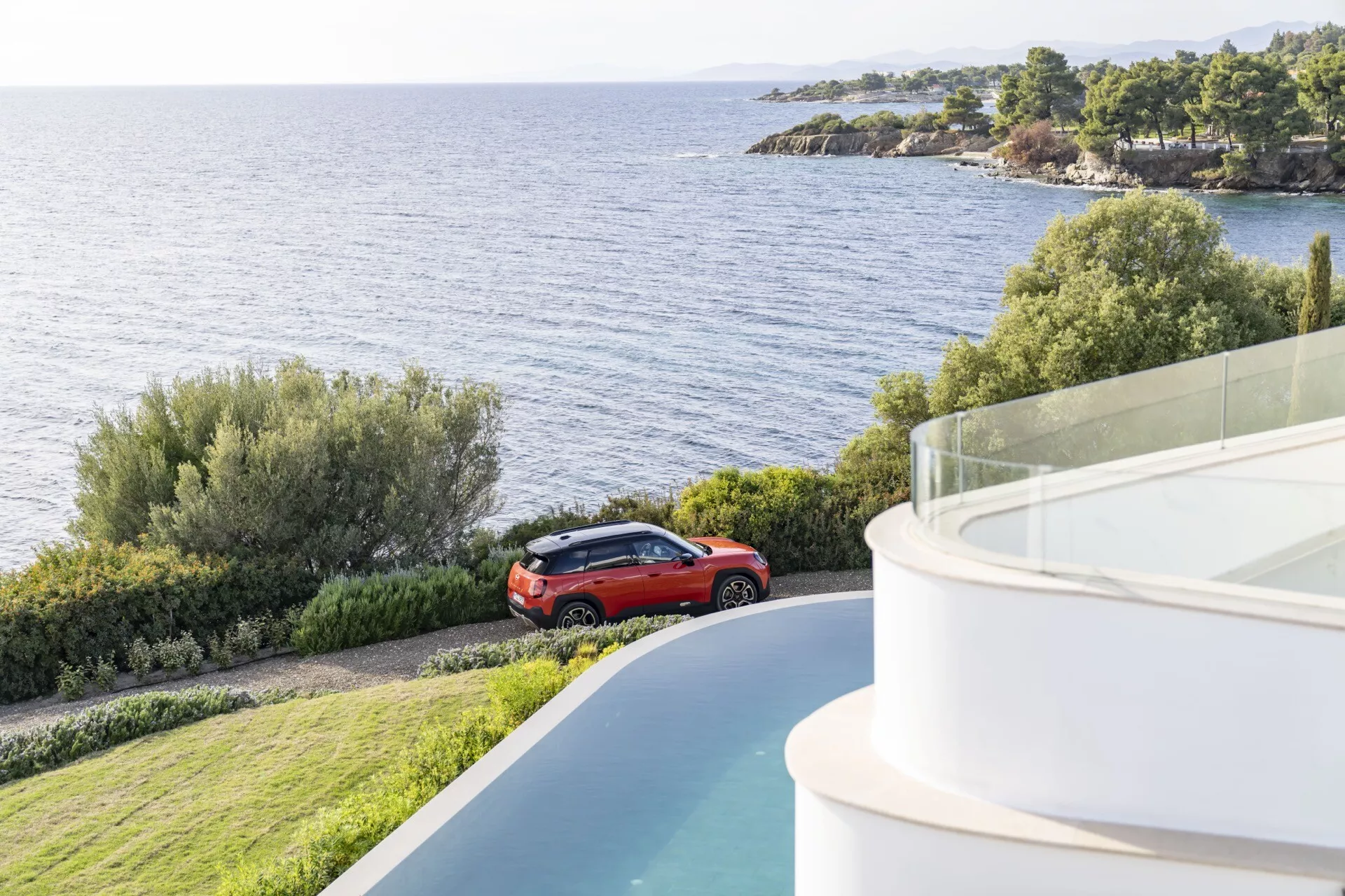 2024 Mini Aceman (J05) in red parked by a scenic coastline or lakeside, viewed from an elevated angle showcasing its crossover design with alloy wheels and a modern, curved architectural building in the foreground