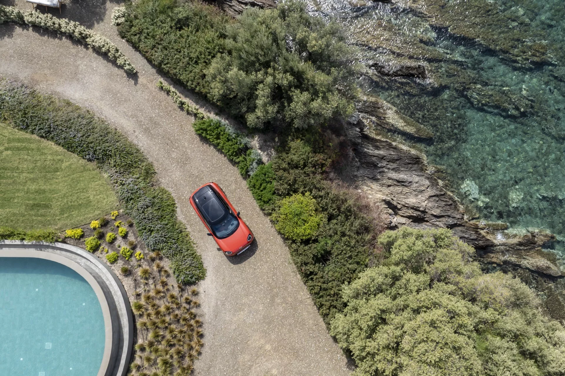 2024 Mini Aceman (J05) in red from a bird's-eye view showcasing its compact hatchback design, sunroof, and side mirrors, positioned on a curved path next to water with lush greenery and part of a swimming pool visible, highlighting its connection to outdoor leisure and adventure.