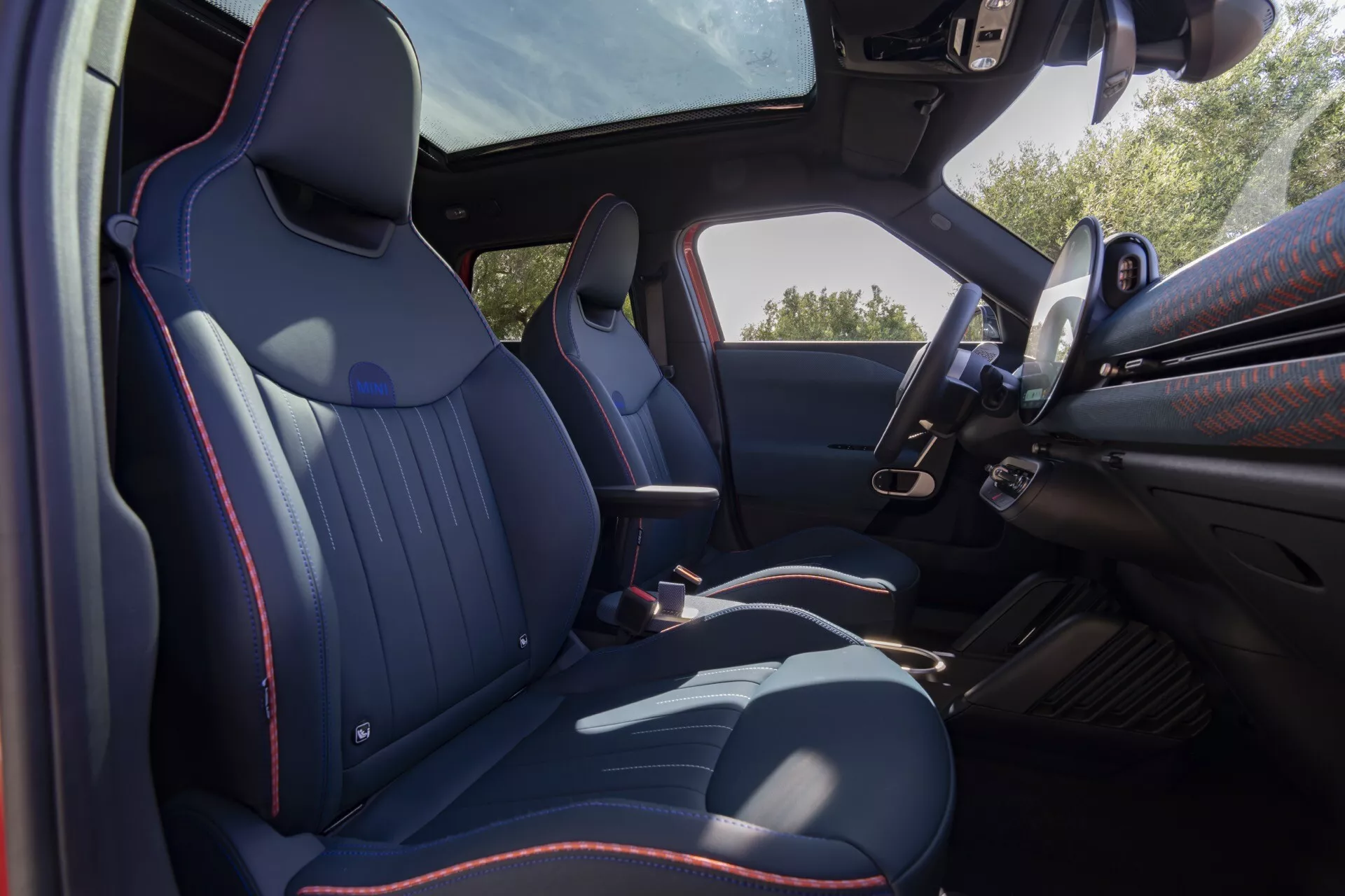 2024 Mini Aceman (JO5) interior view from the rear showing contoured front seats with dark leather and red stitching, driver's door panel, multi-functional steering wheel, center console, panoramic skyroof, and automatic transmission.