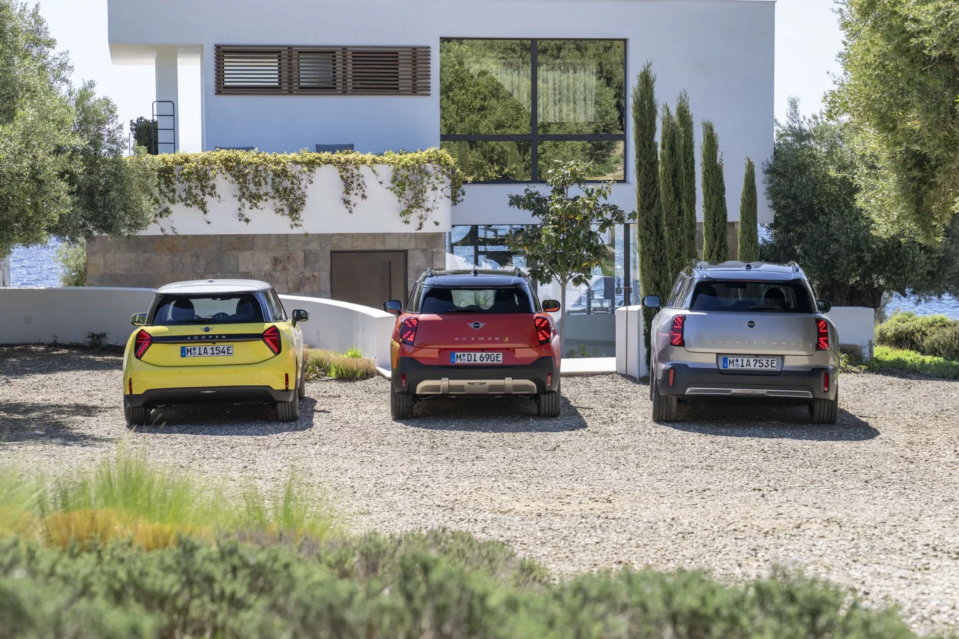 Three 2024 Mini Aceman hatchbacks and SUVs parked in front of a modern architectural building with a clear sky, highlighting their rear designs, LED tail lights, and distinctive colors of yellow, red, and silver.