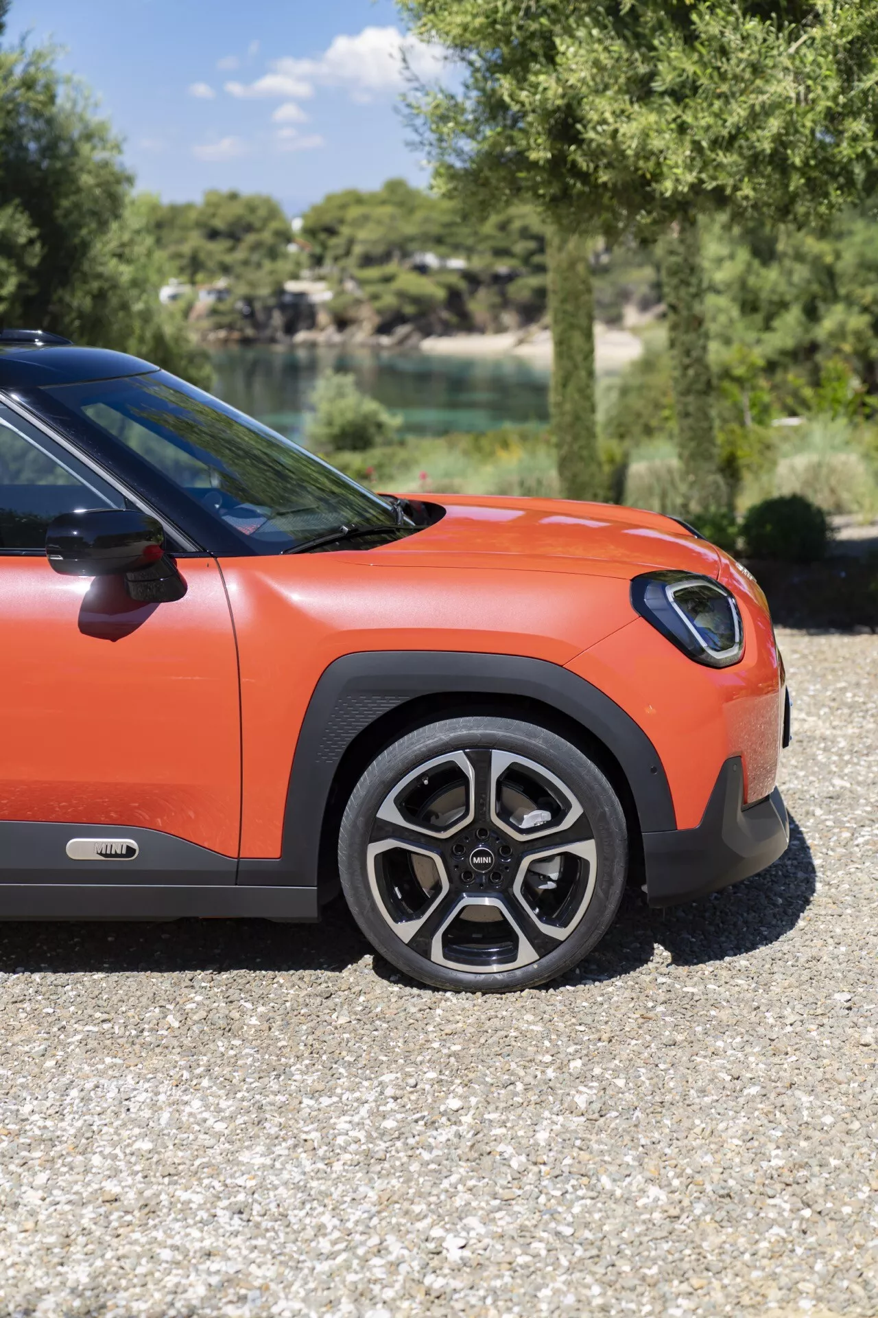 Low angle view of the 2024 Mini Aceman (J05) side front quarter on gravel, featuring an alloy wheel, black over-fender, and bright orange body with a natural lake and trees backdrop.