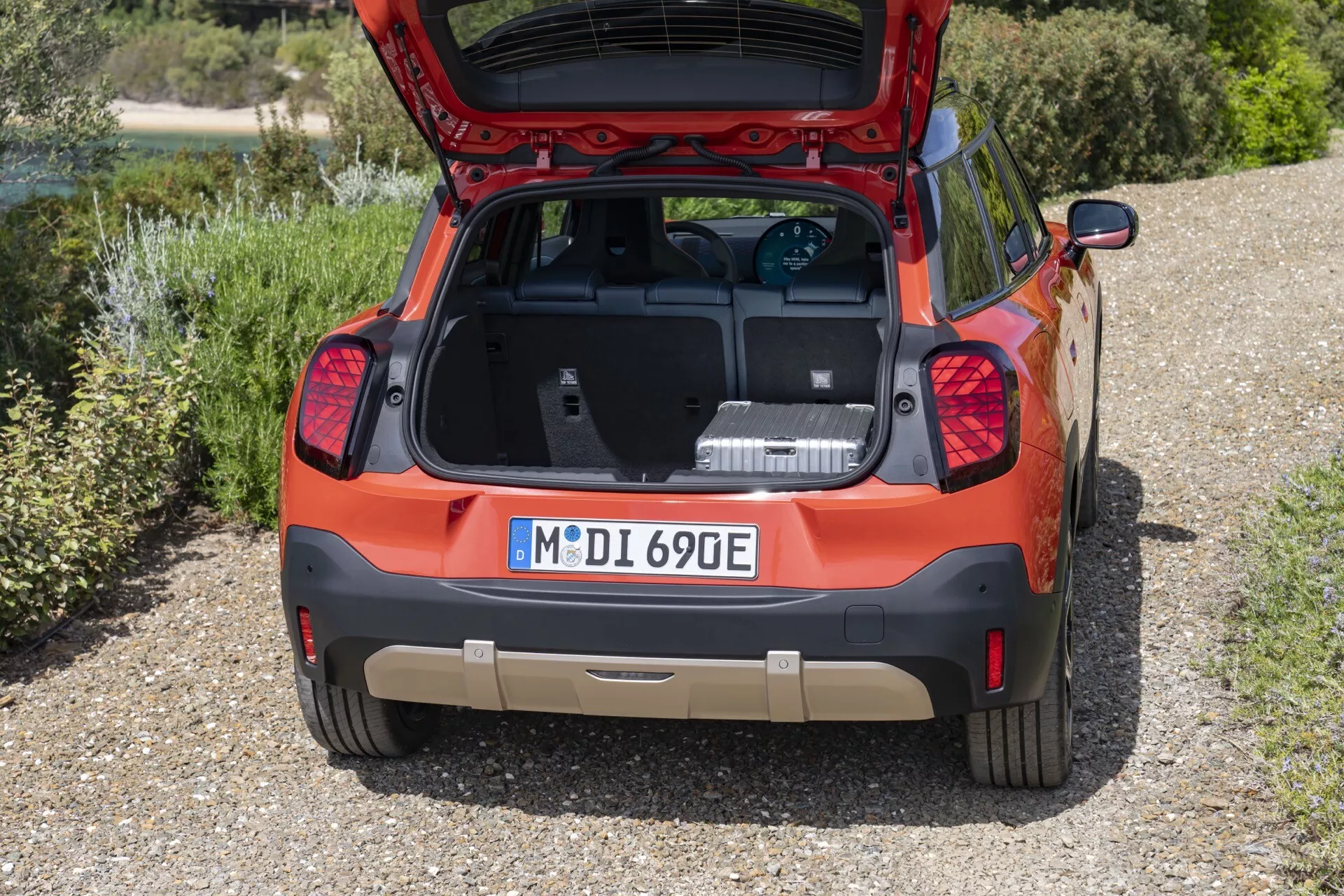 High-angle rear view of a 2024 Mini Aceman (J05) showing open trunk with electric vehicle charging cable, capturing spacious cargo area, design elements and green background.