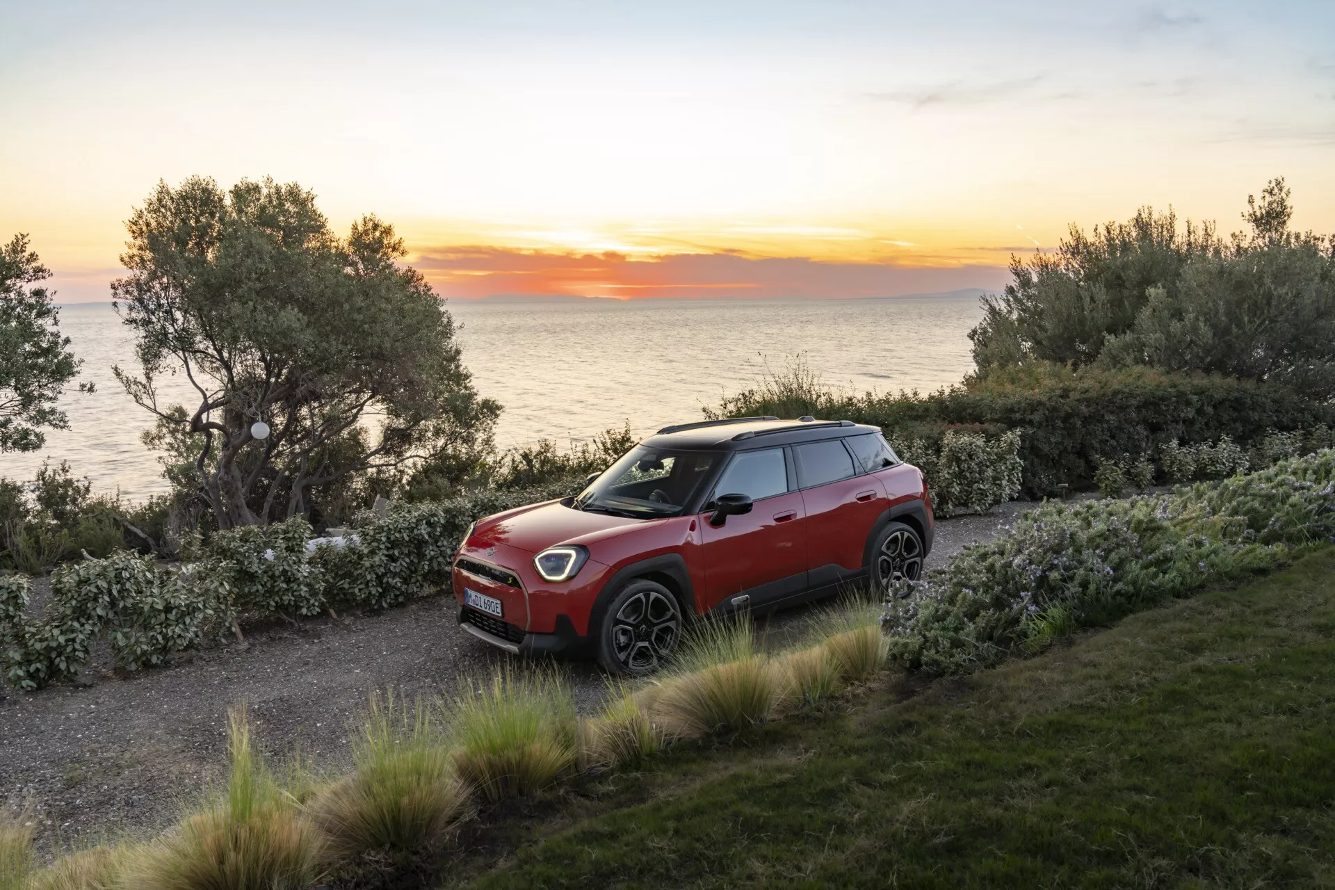 2024 Mini Aceman (J05) three-quarter view on gravel path showcasing front grille, LED headlights, multi-spoke alloy wheels, advanced driving assistance features, two-tone color scheme, compact SUV design, and sunroof in scenic sunrise or sunset setting with trees, shrubbery, and coastal background.