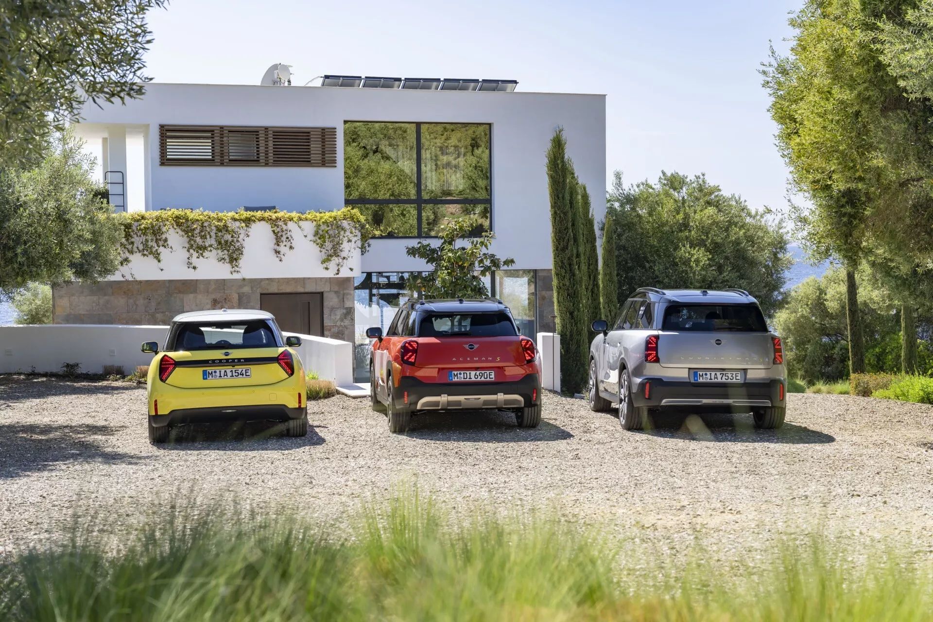 Three Mini Aceman 2024 models (J05) - a yellow convertible, a red hatchback, and a gray SUV - parked in front of a modern two-story house amid lush greenery, showcasing rear three-quarter views and parked parallel on a gravel lot.