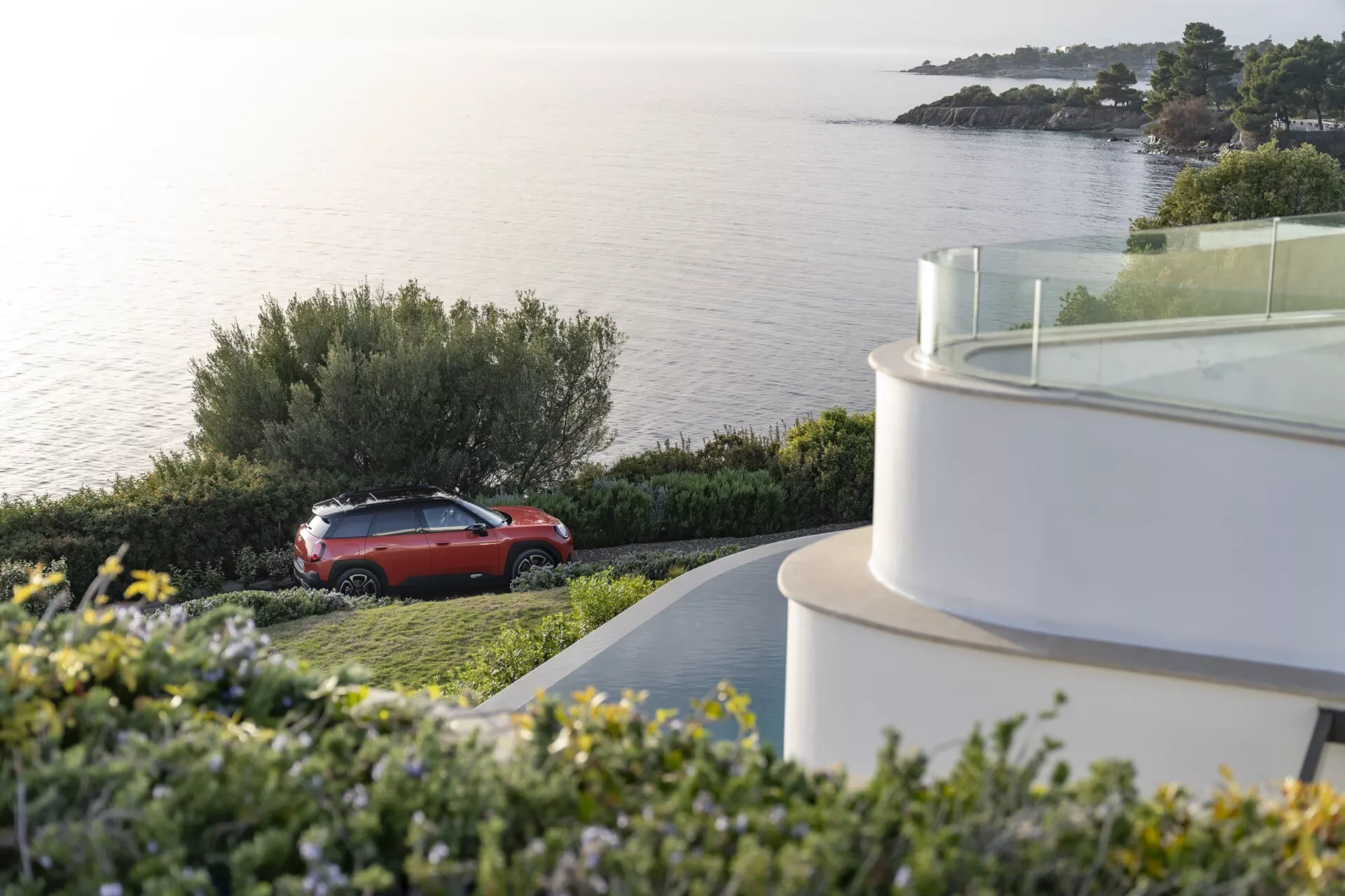 2024 Mini Aceman (J05) in red parked outdoors in a coastal setting, showcasing its SUV side profile, design elements, and alloy wheels against a backdrop of calm sea and natural greenery