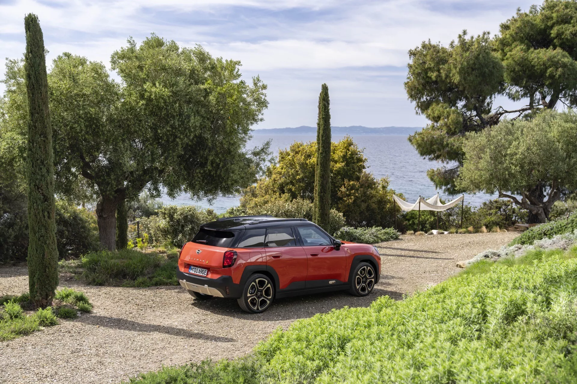 2024 Mini Aceman compact SUV in red, featuring a rear three-quarter view parked on a gravel path in a natural coastal setting with sea and greenery in the background, highlighting rear lights, exhaust pipes, hatch door with wiper, contrasting roof color, and alloy wheels.