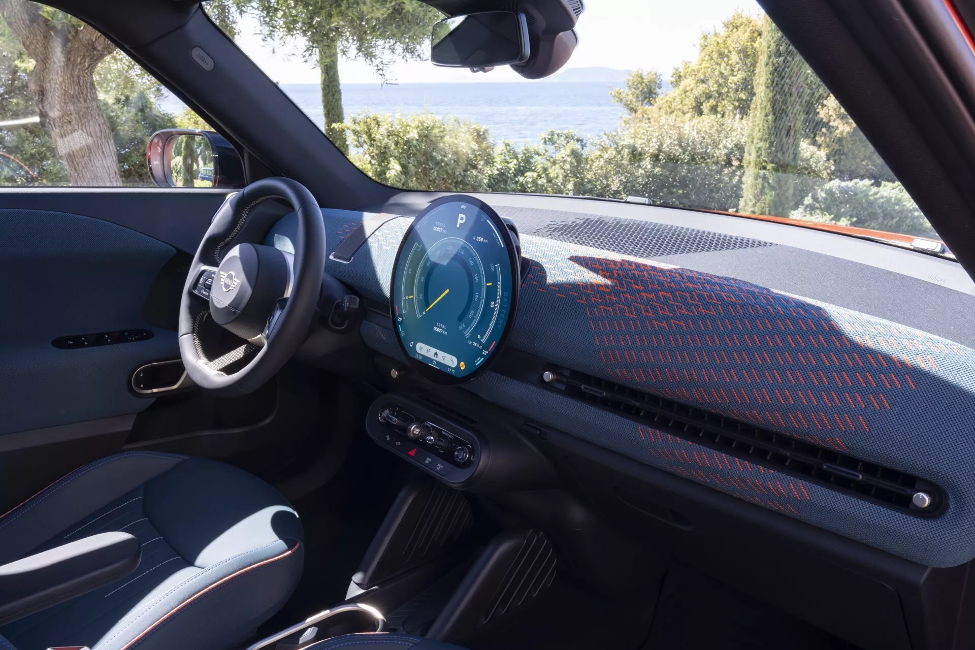 Interior view of the Mini Aceman 2024 (JO5) highlighting the driver's area with a modern dashboard, digital instrument cluster, and advanced steering wheel controls, parked in a natural setting