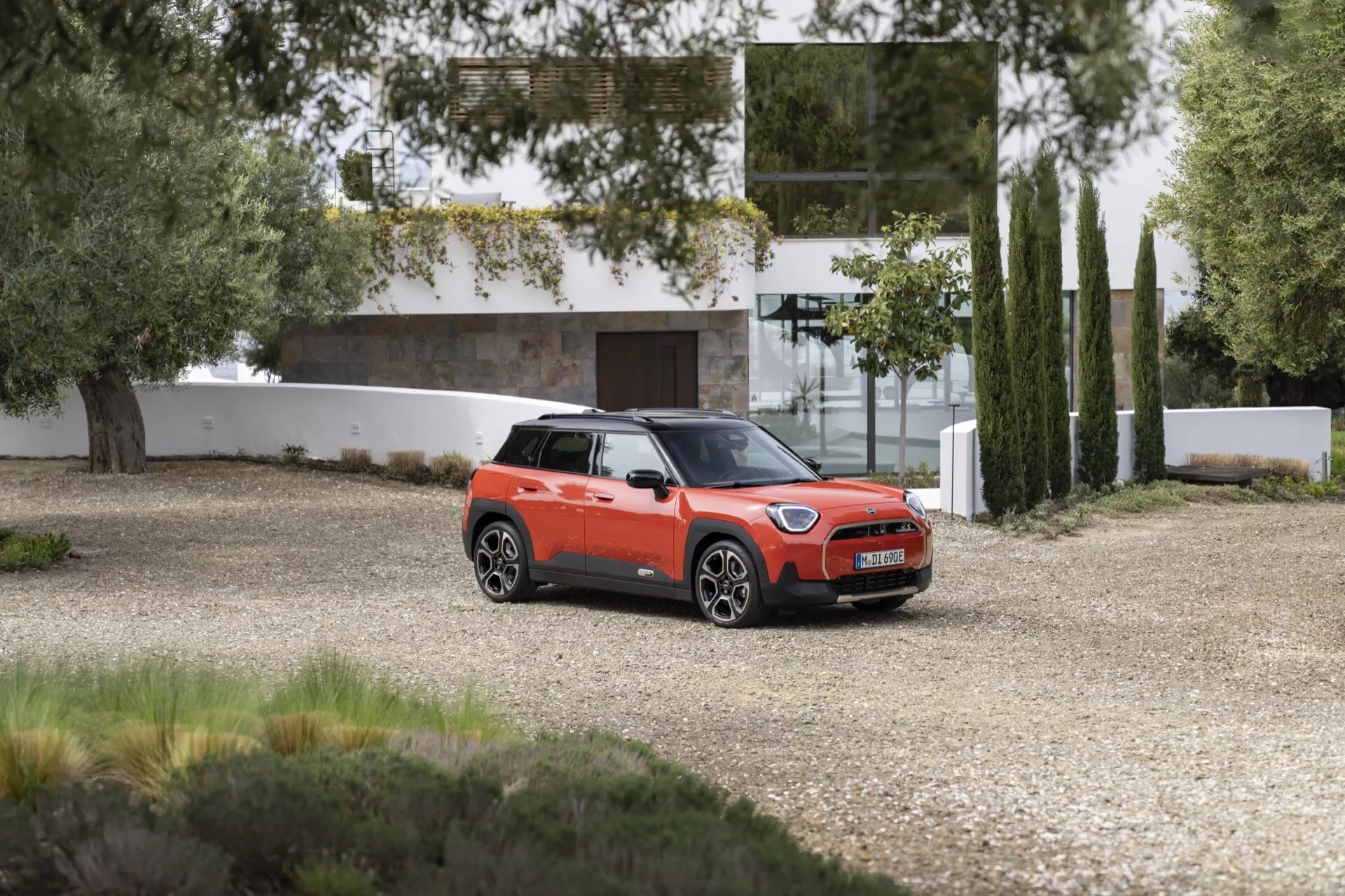 2024 Mini Aceman compact SUV in vibrant orange with black roof, parked on gravel showing a three-quarter front view against a residential backdrop