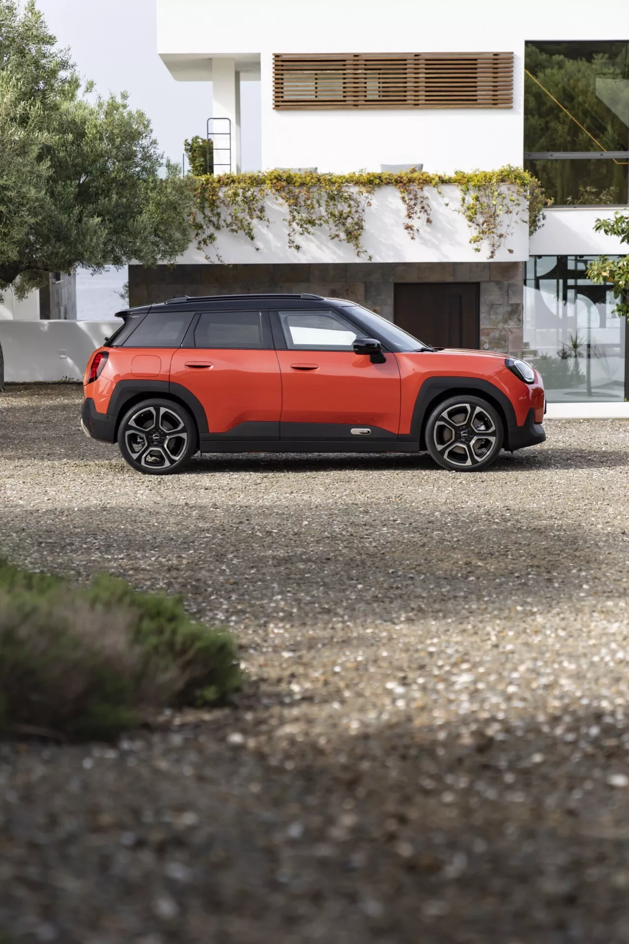 2024 Mini Aceman (J05) compact SUV side profile view on gravel, featuring raised clearance, prominent wheel arches, roof rails, modern black trim windows, two-tone orange and black body, protective door molding, and alloy wheels