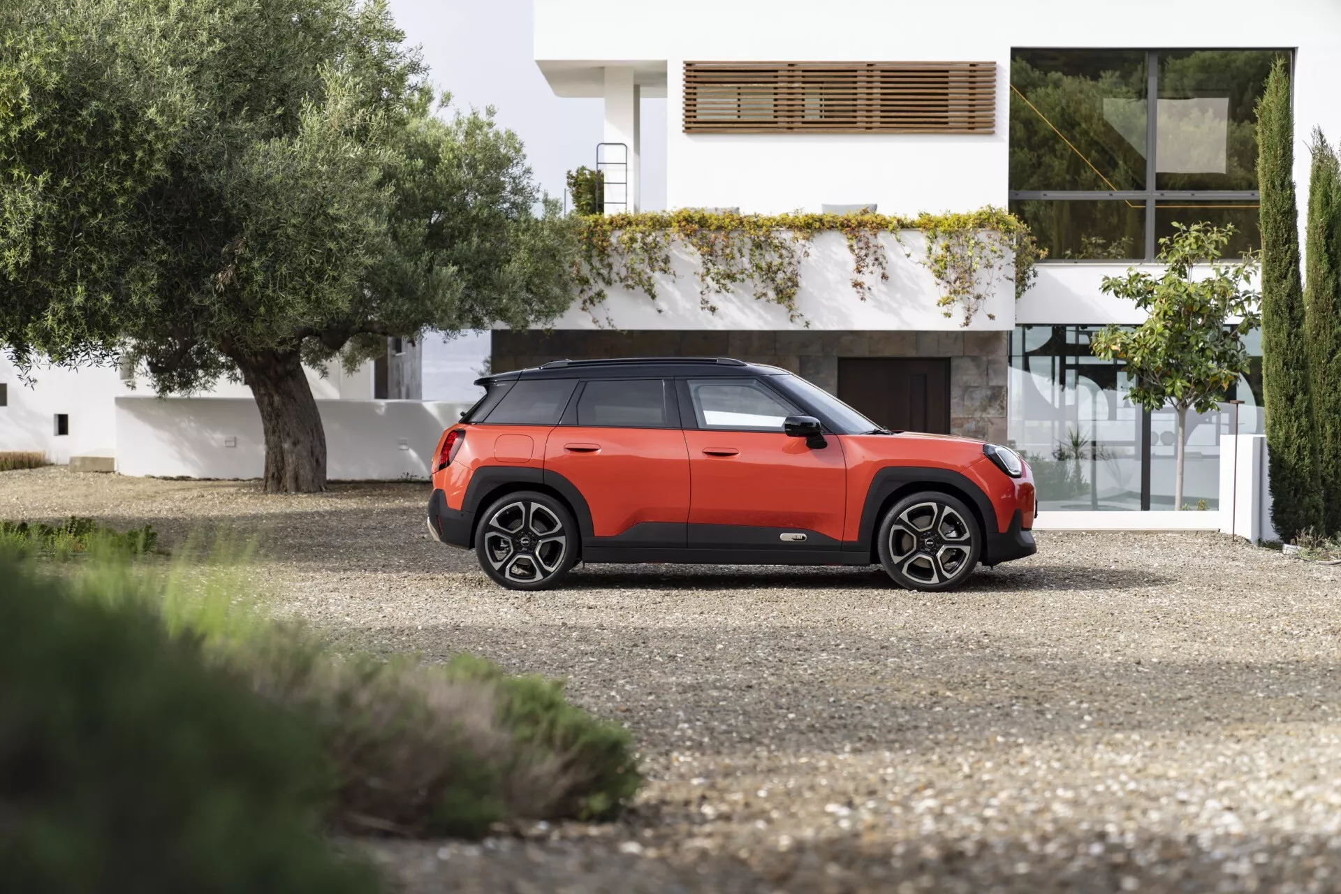 2024 Mini Aceman SUV in a bold orange-red and black color scheme parked in front of a contemporary house, showcasing its front and side profiles with distinctive design lines, LED headlamps, multi-spoke wheels, and high ground clearance.