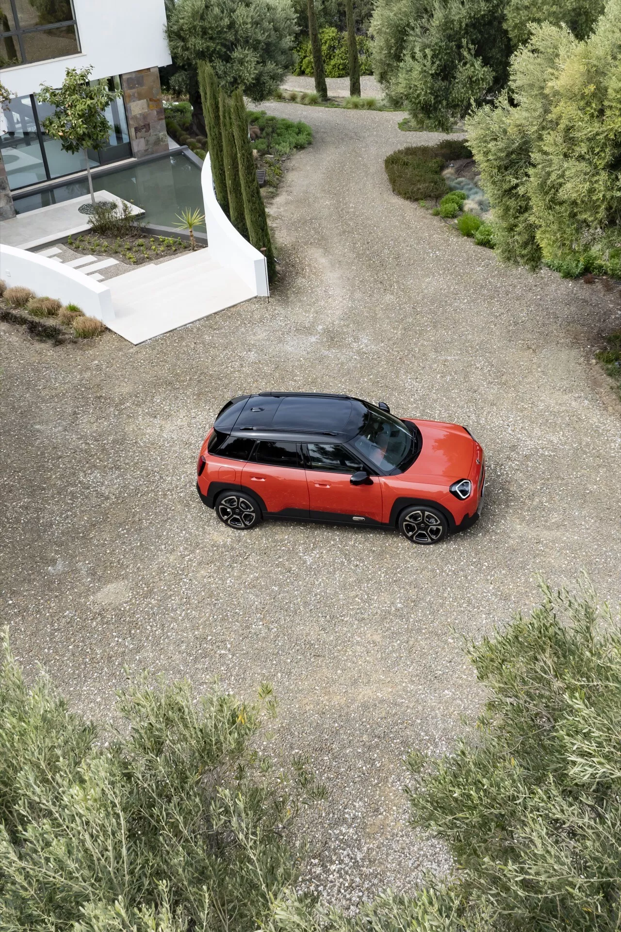 Overhead view of 2024 Mini Aceman (J05) in bright red with black roof parked on gravel path leading to modern building amongst greenery, showcasing compact SUV design, two-tone paint, panoramic roof, and multi-spoke alloy wheels.