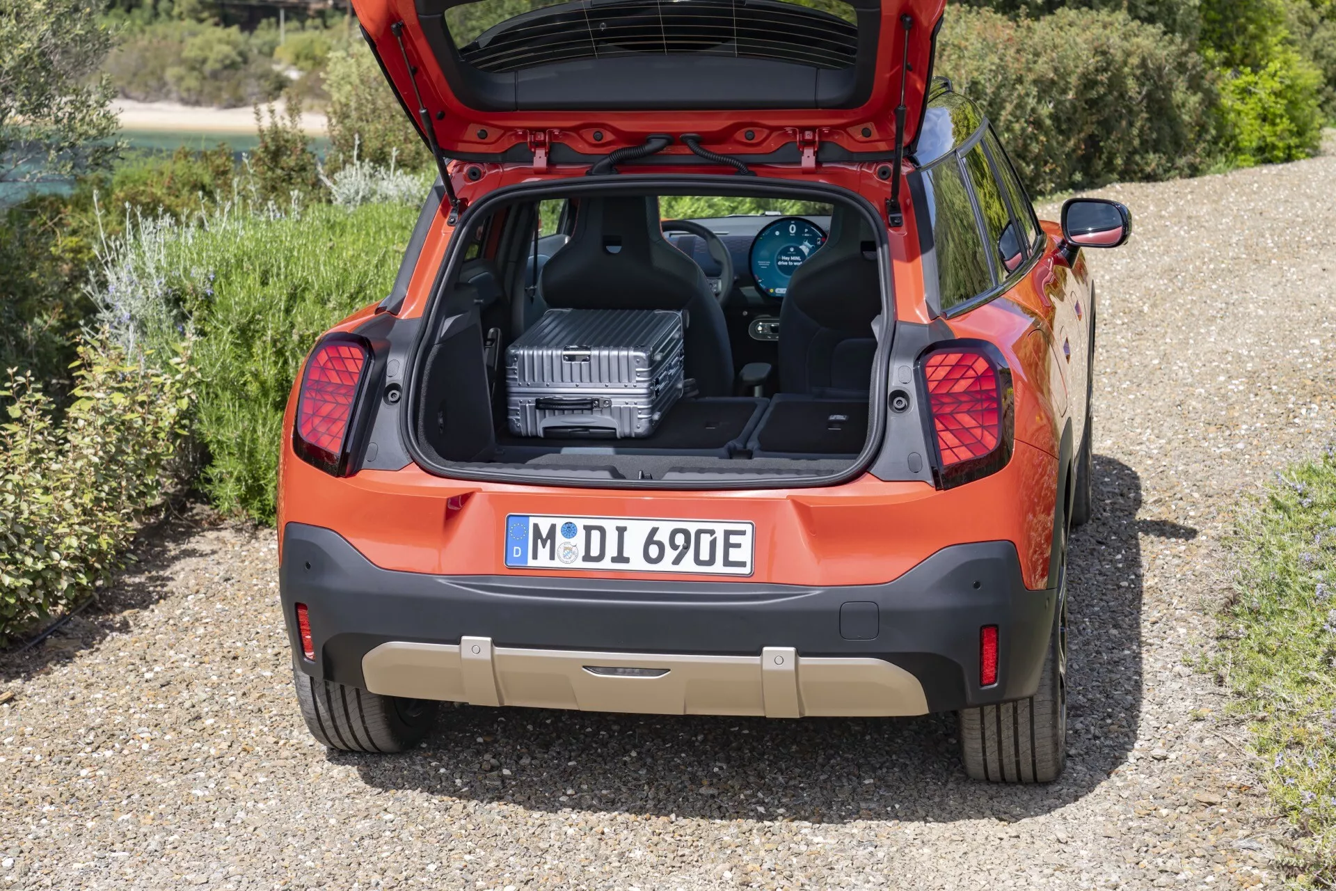 Rear view of an orange 2024 Mini Aceman (J05) hatchback, distinguished by its electric or hybrid powertrain, with an open trunk displaying a large powertrain component, possibly for educational or service purposes, parked on a gravel surface surrounded by greenery.