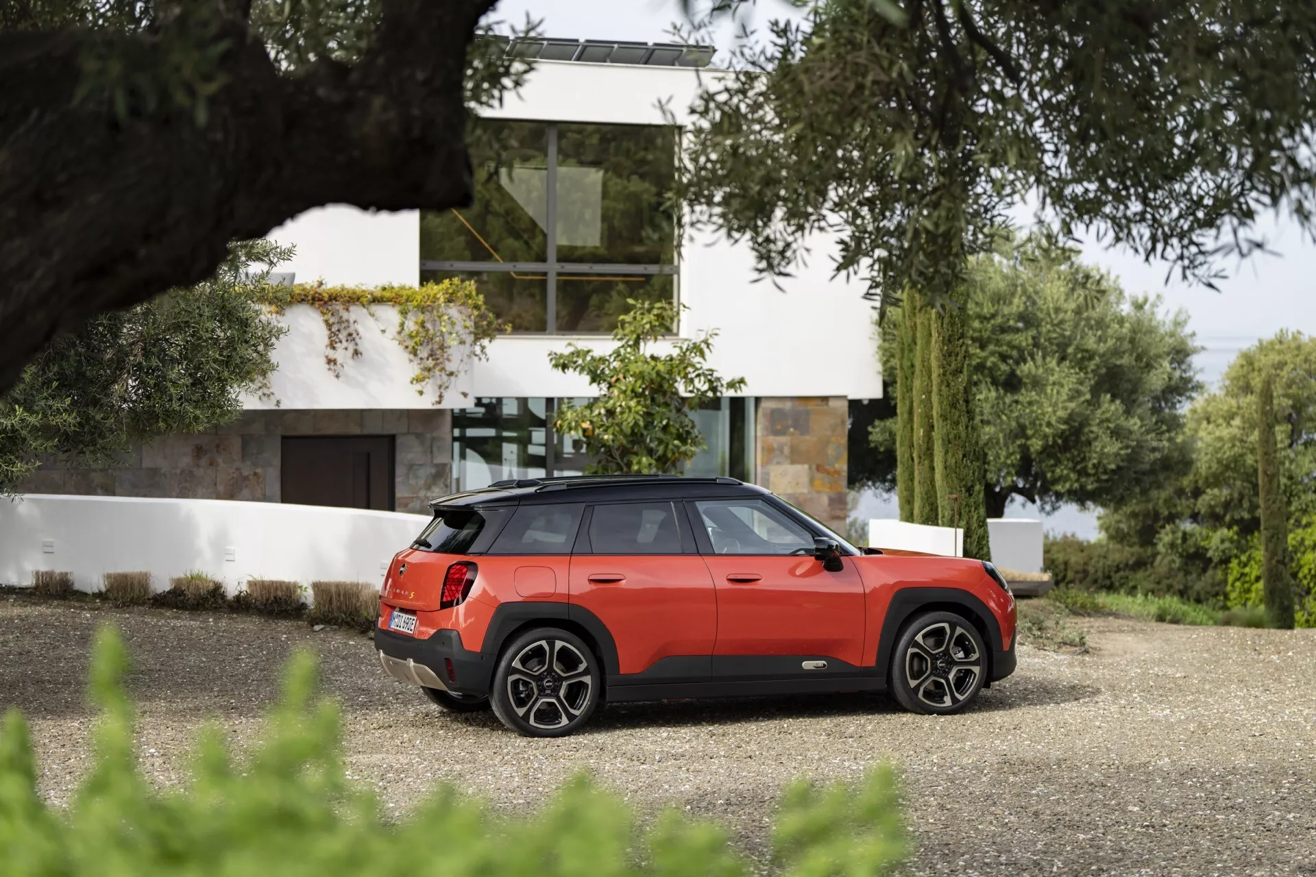2024 Mini Aceman (J05) in metallic red parked on a driveway, showcasing its sporty and luxurious design with a panoramic sunroof, alloy wheels, rear LED lighting, and advanced safety features in a contemporary residential setting