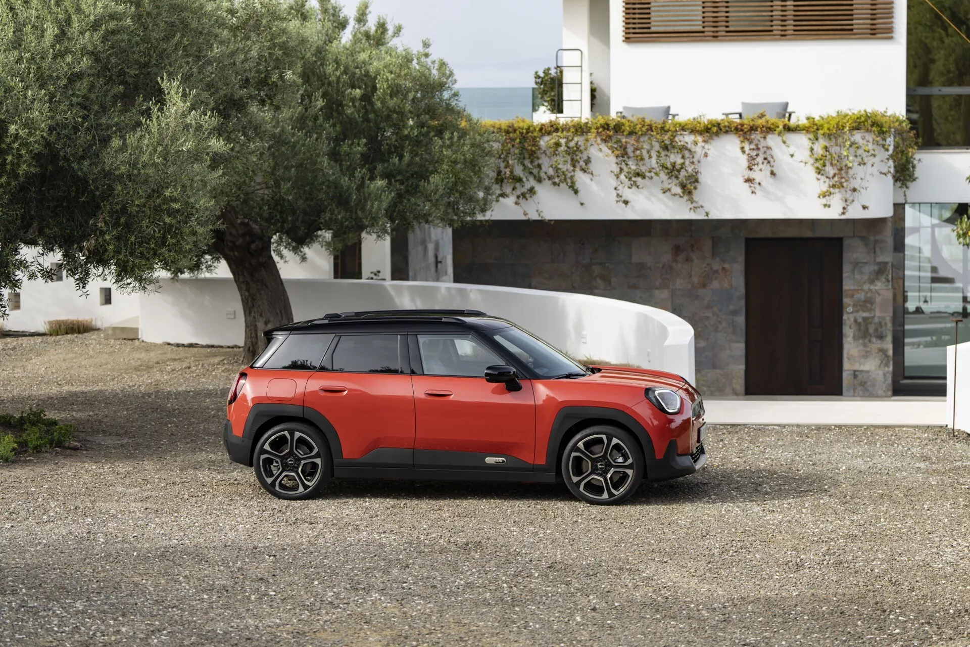 2024 Mini Aceman (J05) in red with black roof, showcasing its compact SUV design from a three-quarter front angle on a gravel surface, highlighting the front grille, sleek headlights, sporty wheels, and distinctive side profile against a contemporary residential backdrop