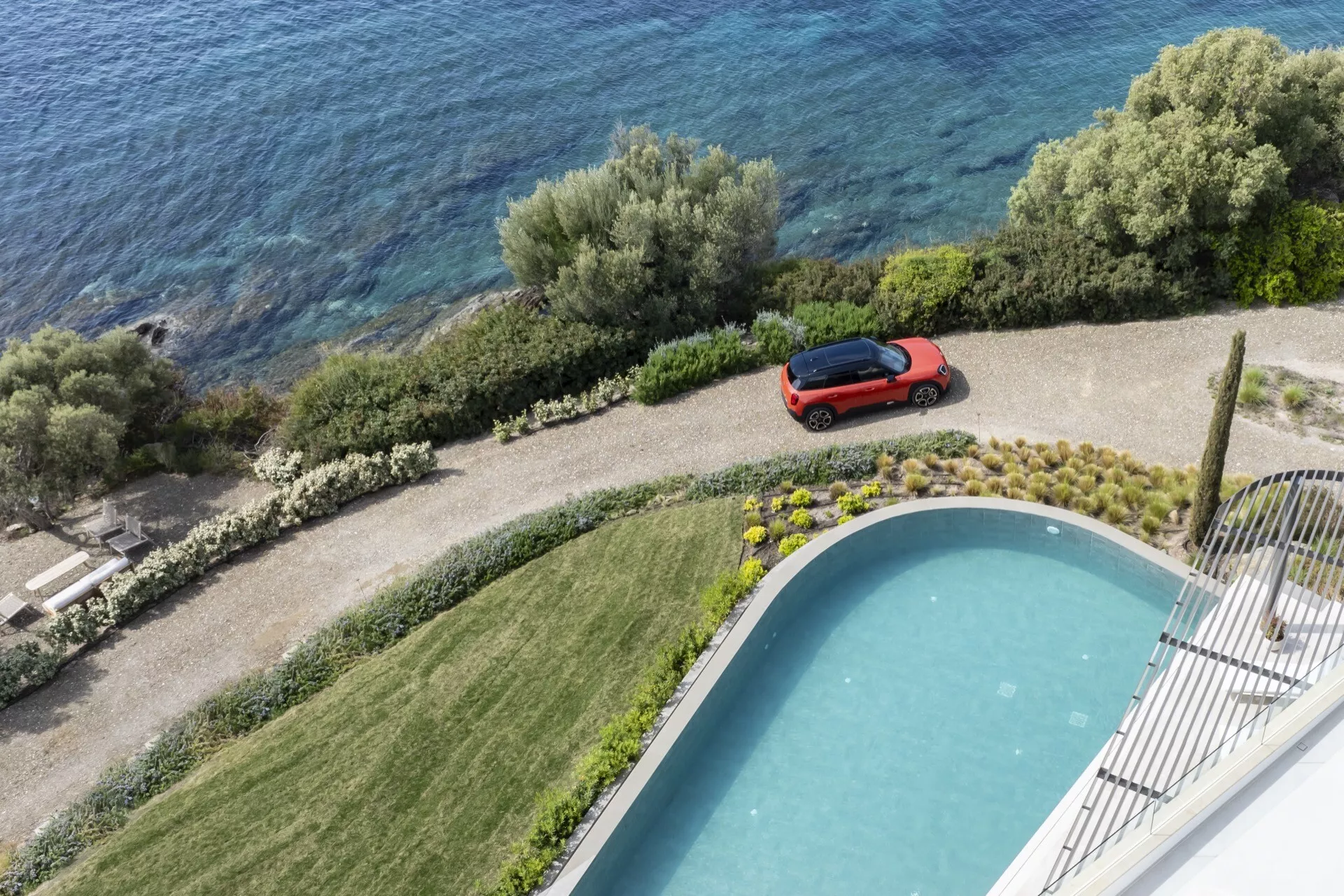 2024 Mini Aceman (J05) in red, parked at a luxurious estate with a view of a pool and waterfront, highlighting its aerodynamic design and glossy finish